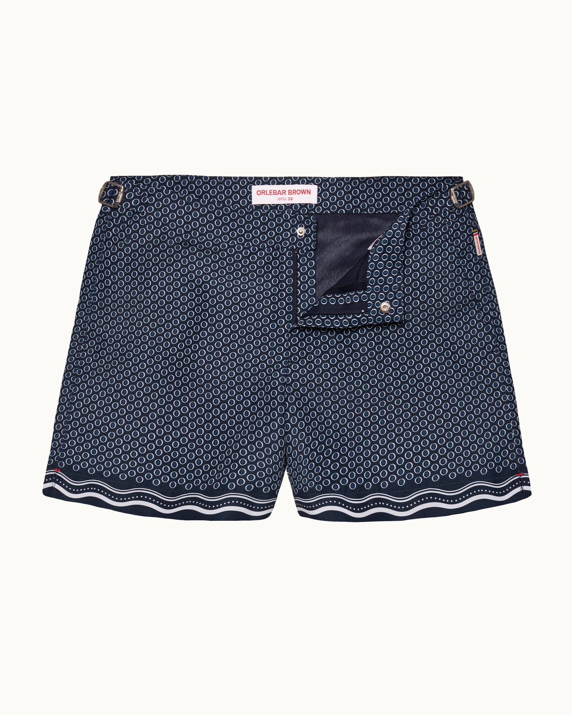 A pair of men's swimshorts featuring a navy blue and white geometric pattern, a solid navy back pocket, and a wavy white stripe along the hem, displayed against a light cream background.
