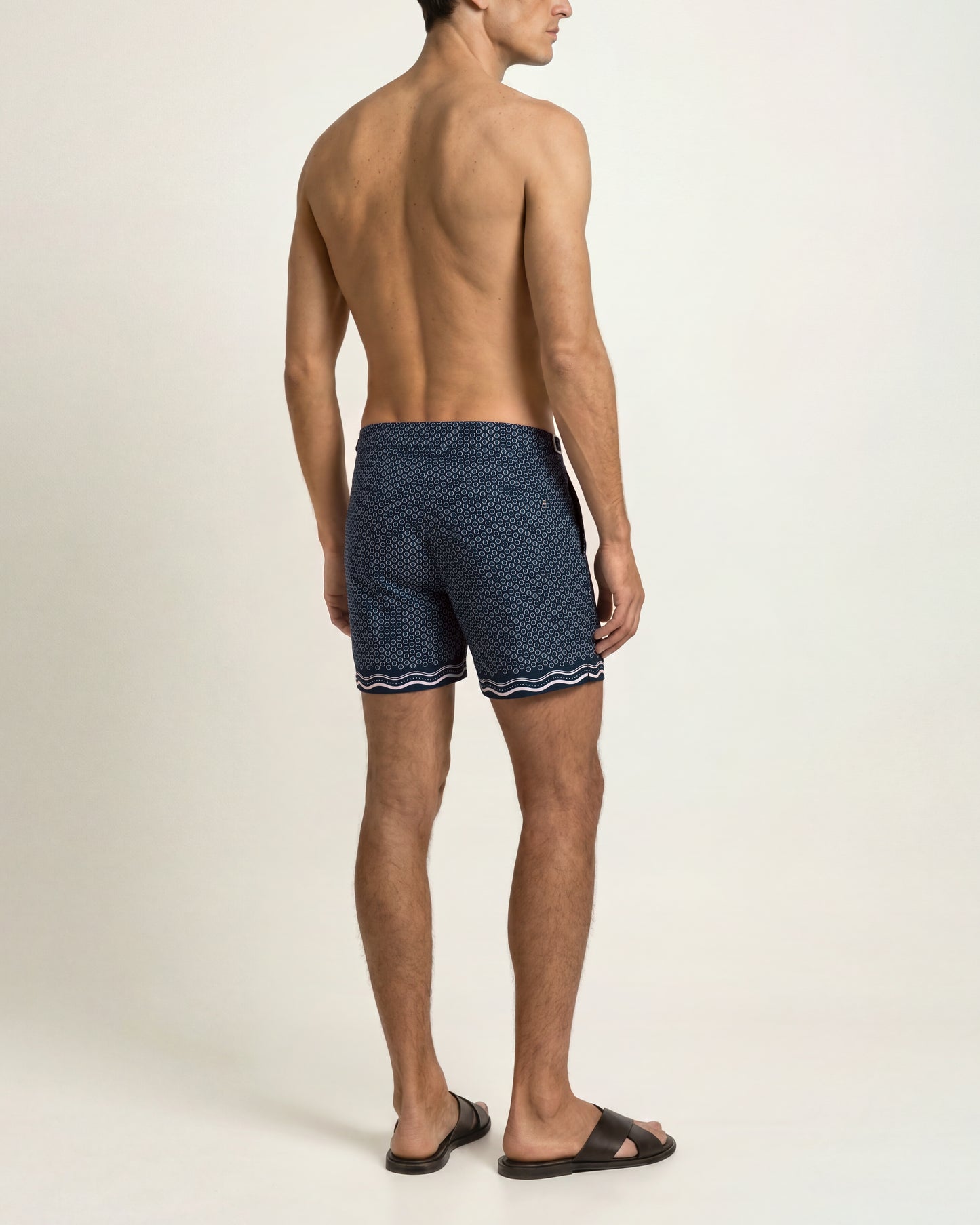 A man shown from the back wearing navy blue patterned swim trunks with a white wavy trim, standing against a plain light beige studio background.