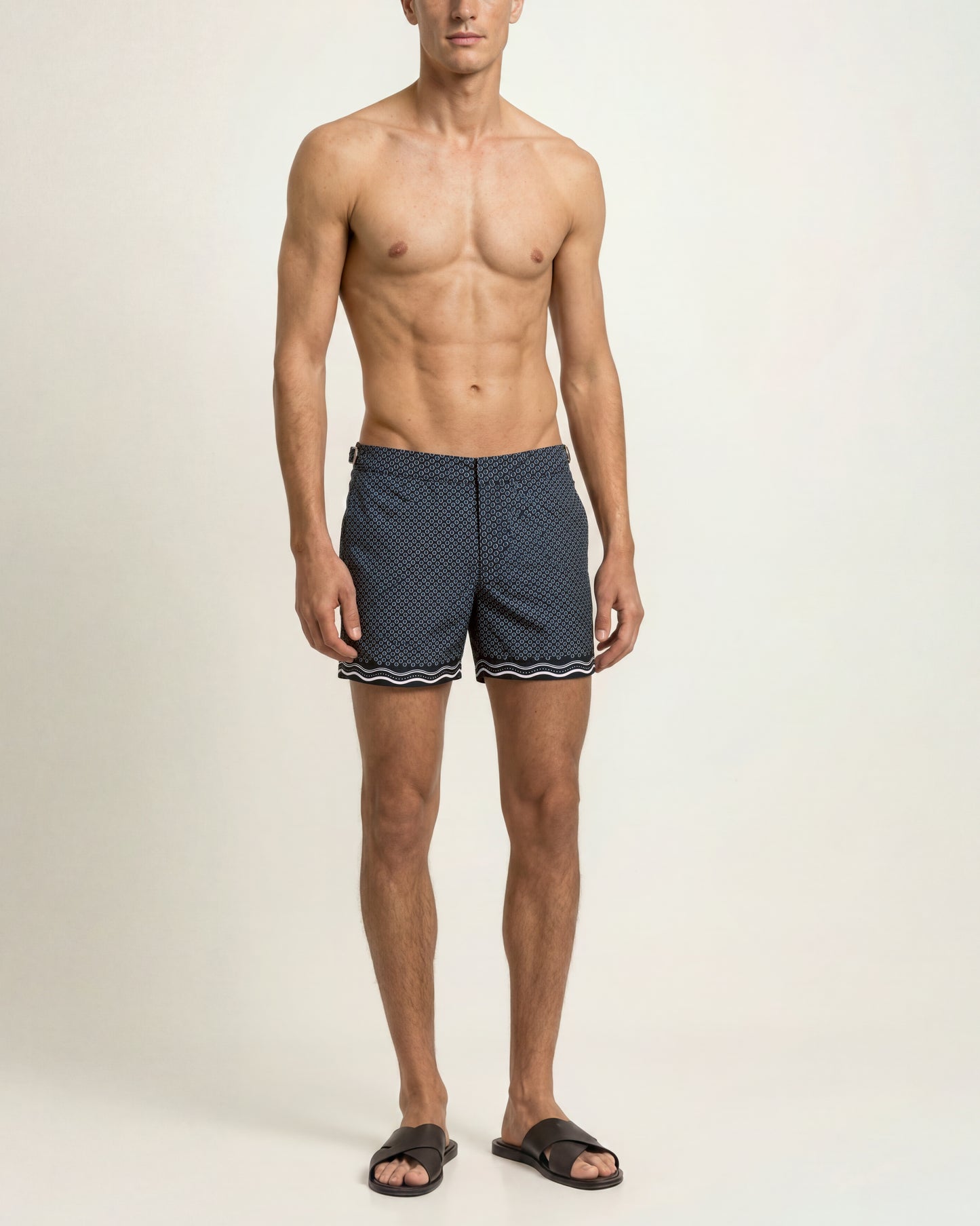 A shirtless male model wearing navy blue patterned swim trunks with a white wavy trim and black slide sandals, standing against a plain off-white studio background.