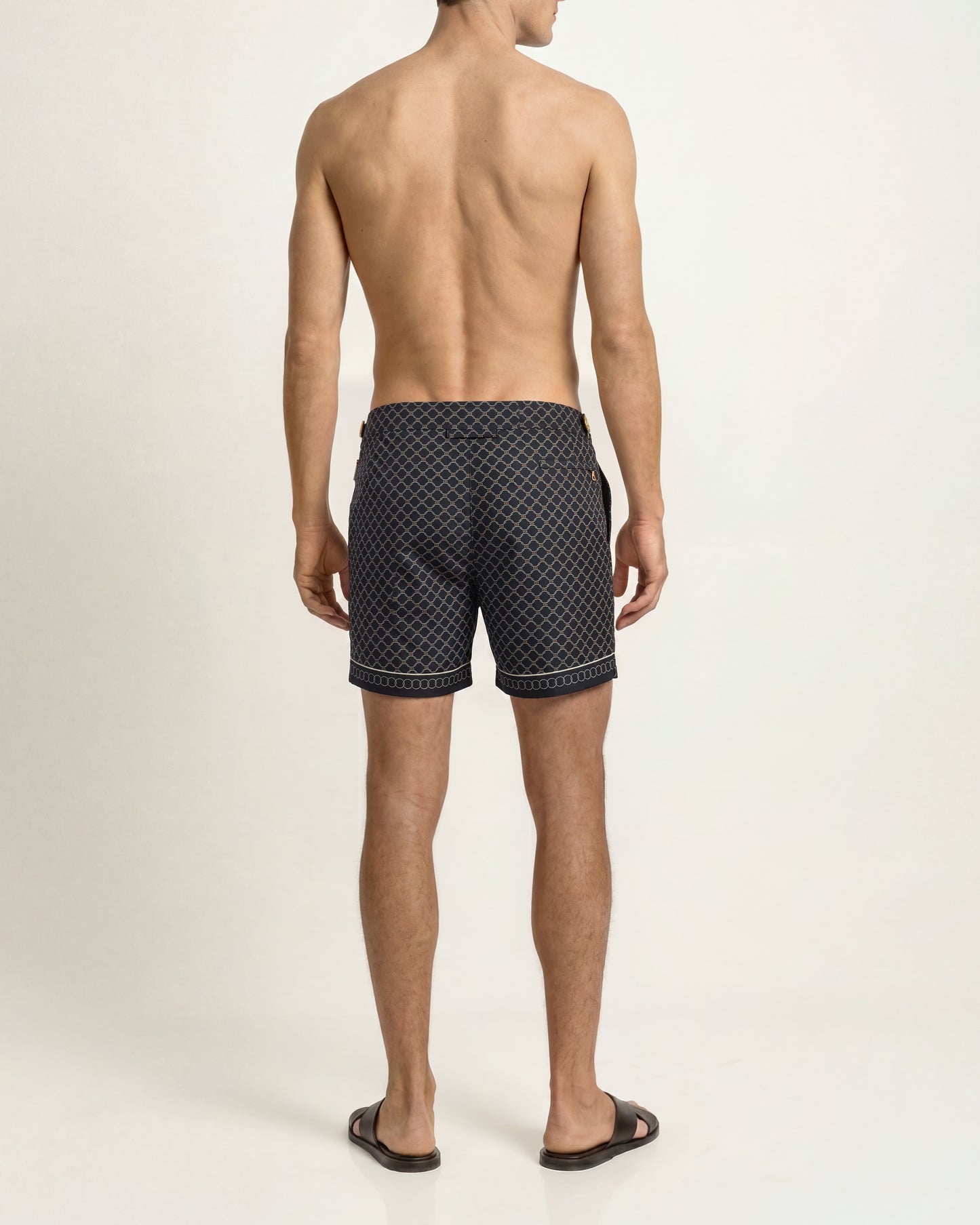 Rear view of a man wearing dark patterned swim trunks and black slide sandals against a plain light background.