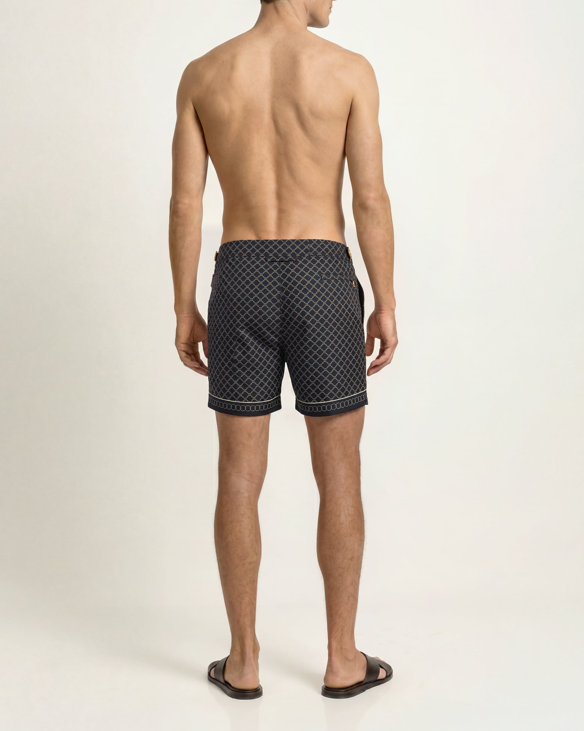Rear view of a man wearing dark patterned swim trunks and black slide sandals against a plain light background.
