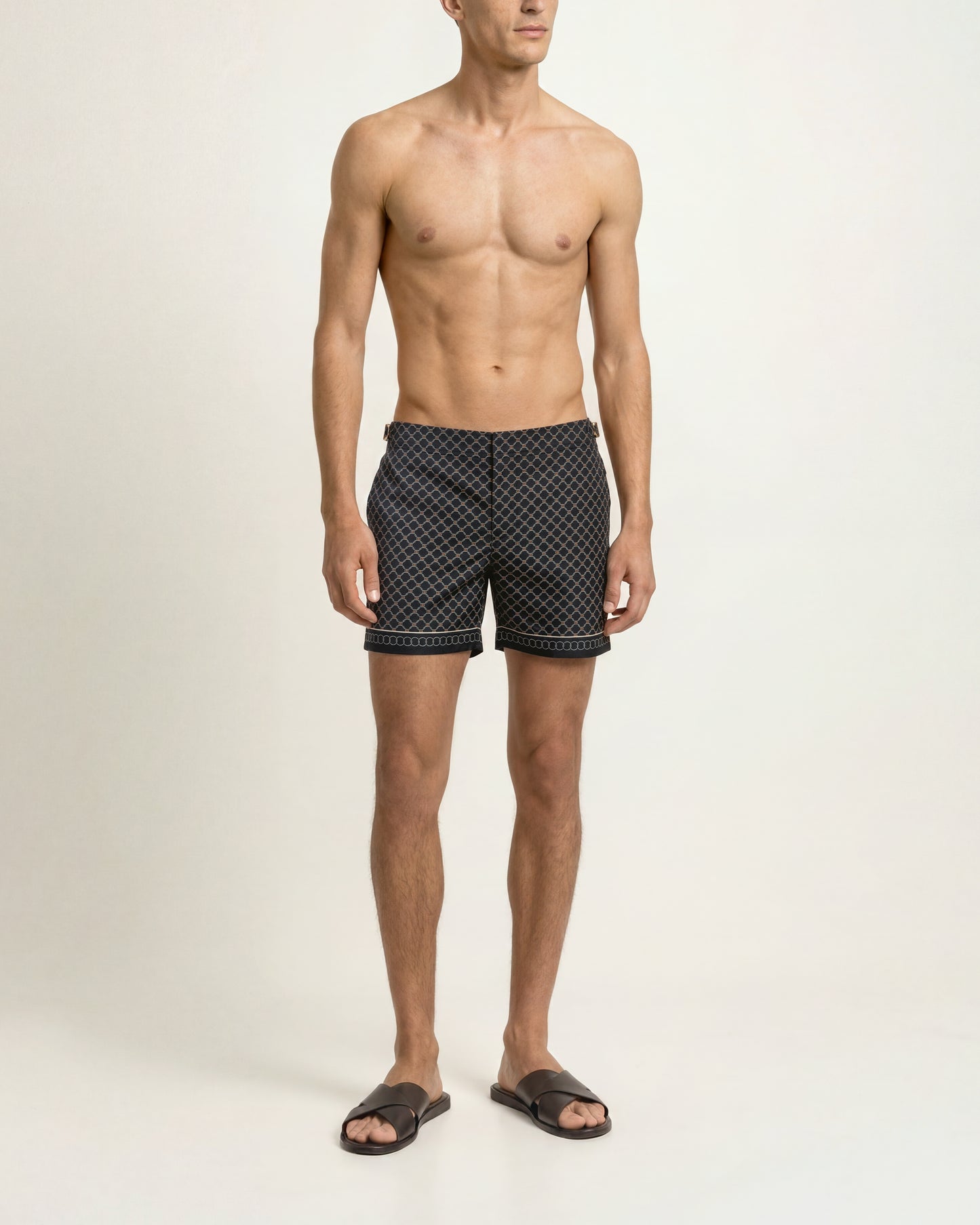 A shirtless male model wearing black swim trunks with a small white geometric pattern, standing against a plain off-white studio background.
