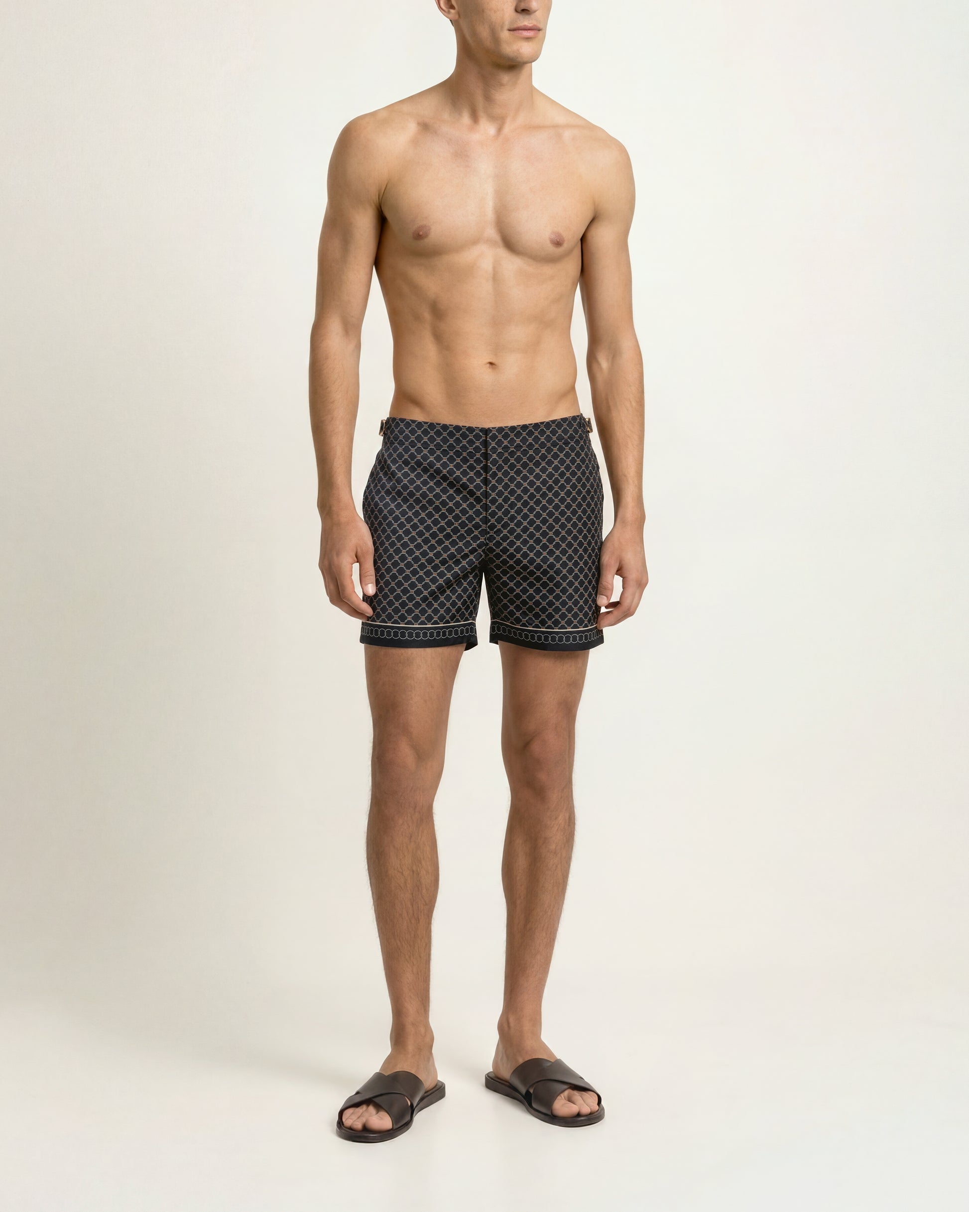 A shirtless male model wearing black swim trunks with a small white geometric pattern, standing against a plain off-white studio background.