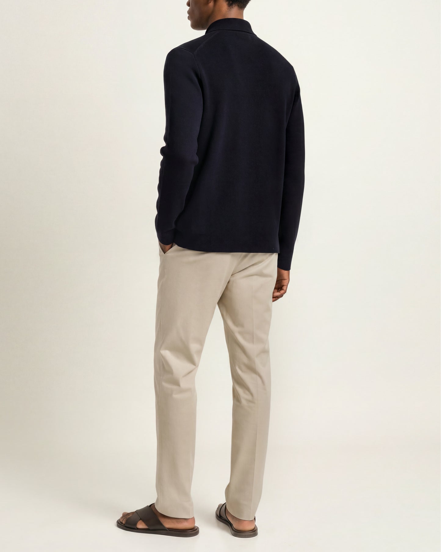 A man shown from the back wearing a dark navy long-sleeve knit top, beige trousers, and dark brown sandals against a plain off-white studio background.