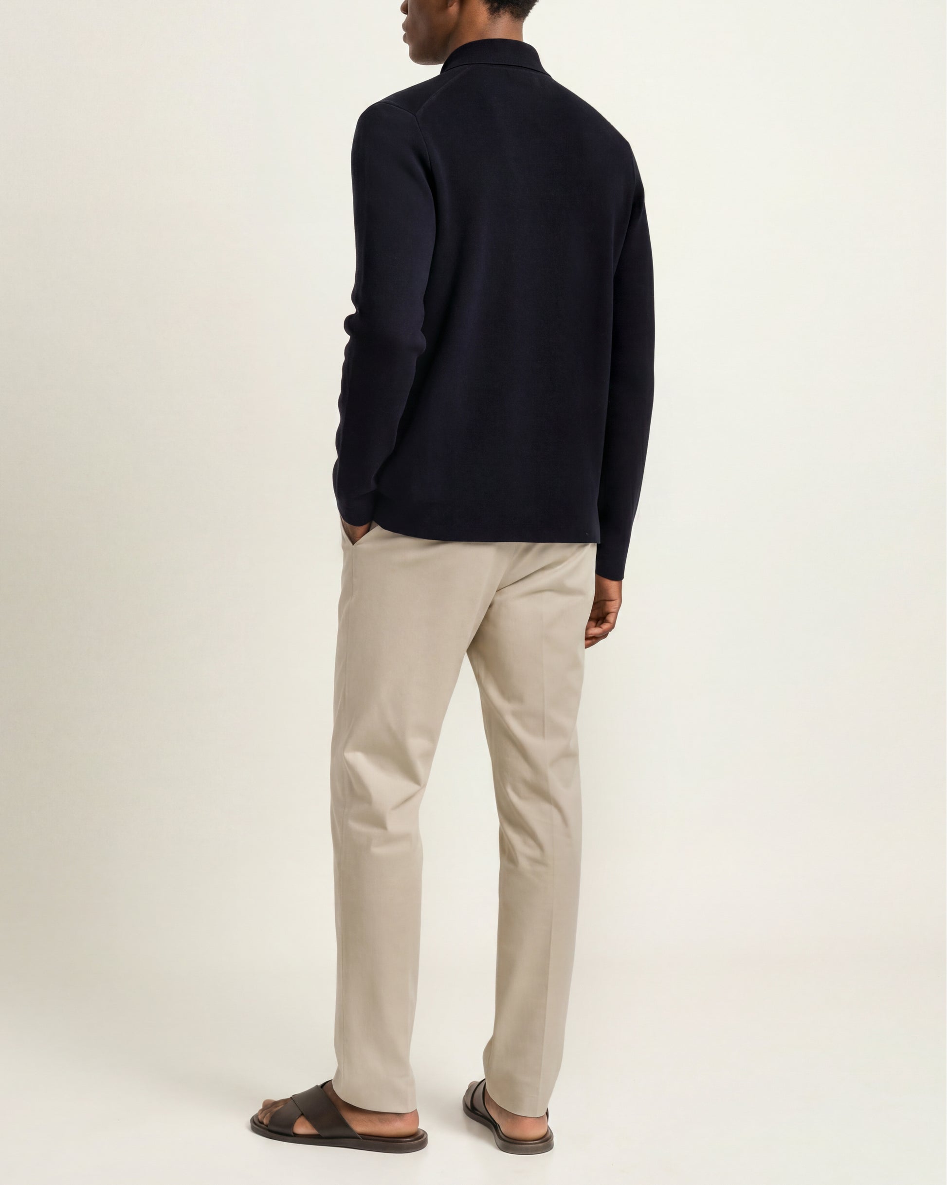 A man shown from the back wearing a dark navy long-sleeve knit top, beige trousers, and dark brown sandals against a plain off-white studio background.