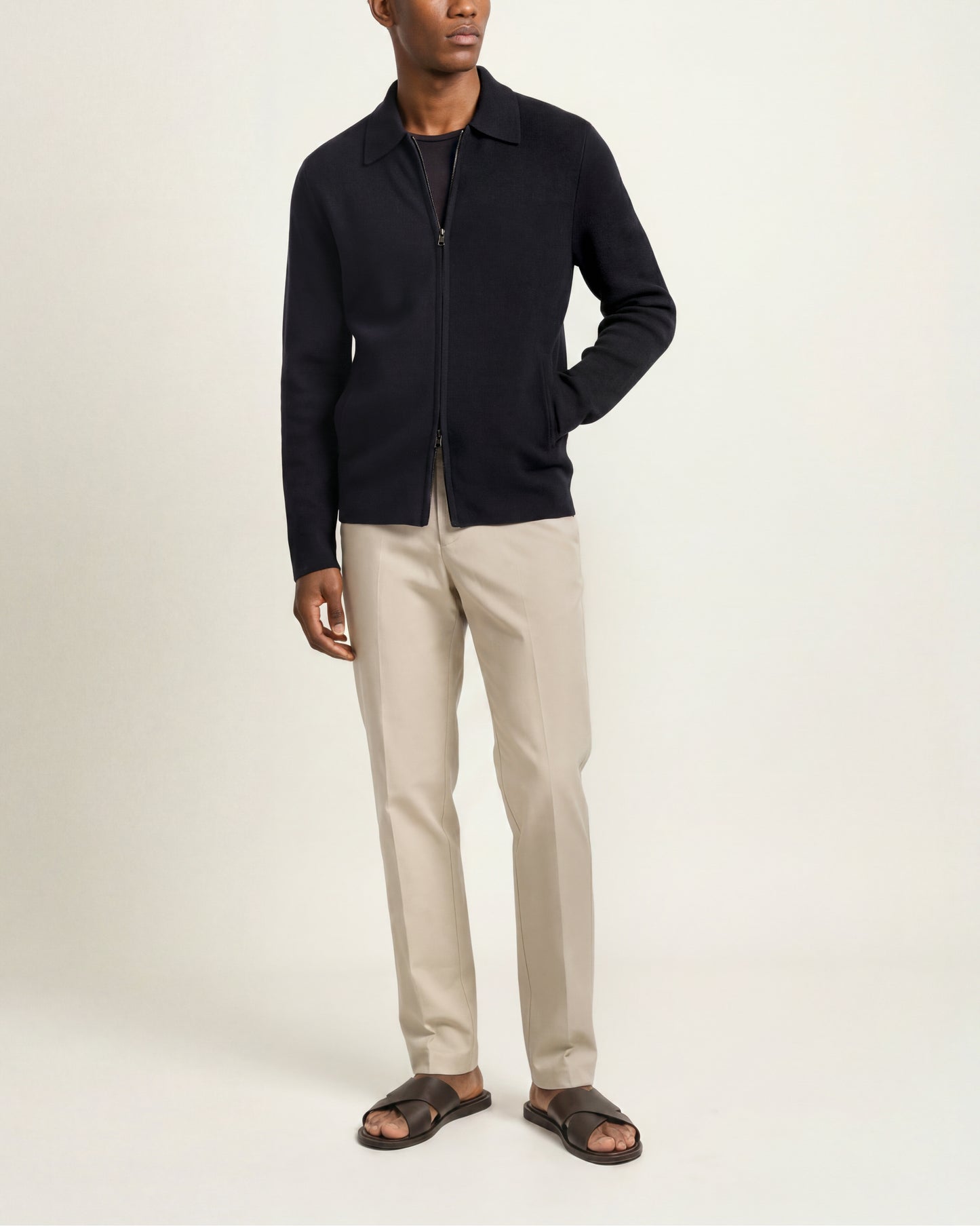 A man modeling a black zip-up knit cardigan over a black shirt, paired with beige trousers and dark brown slide sandals, against a plain off-white studio background.