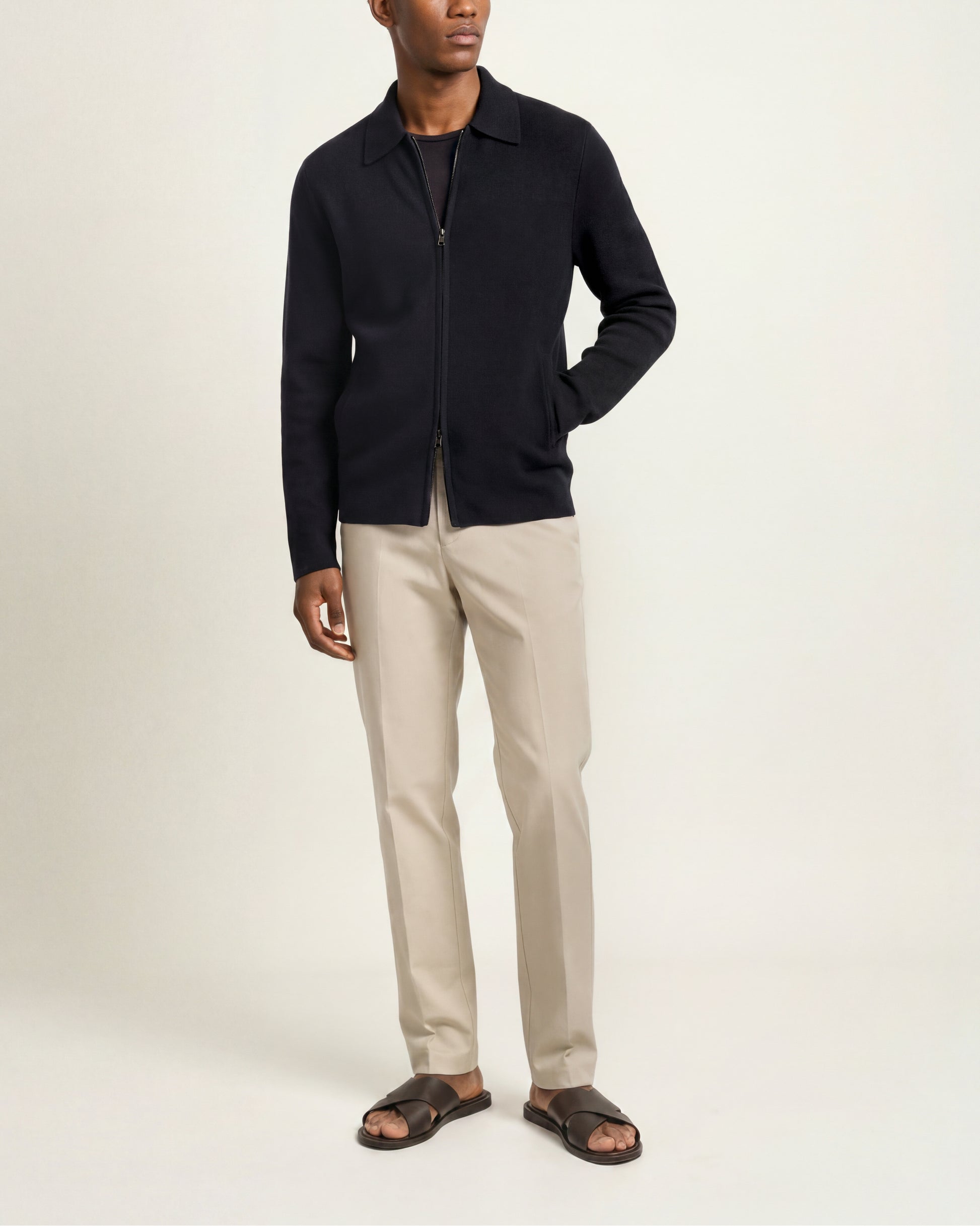 A man modeling a black zip-up knit cardigan over a black shirt, paired with beige trousers and dark brown slide sandals, against a plain off-white studio background.