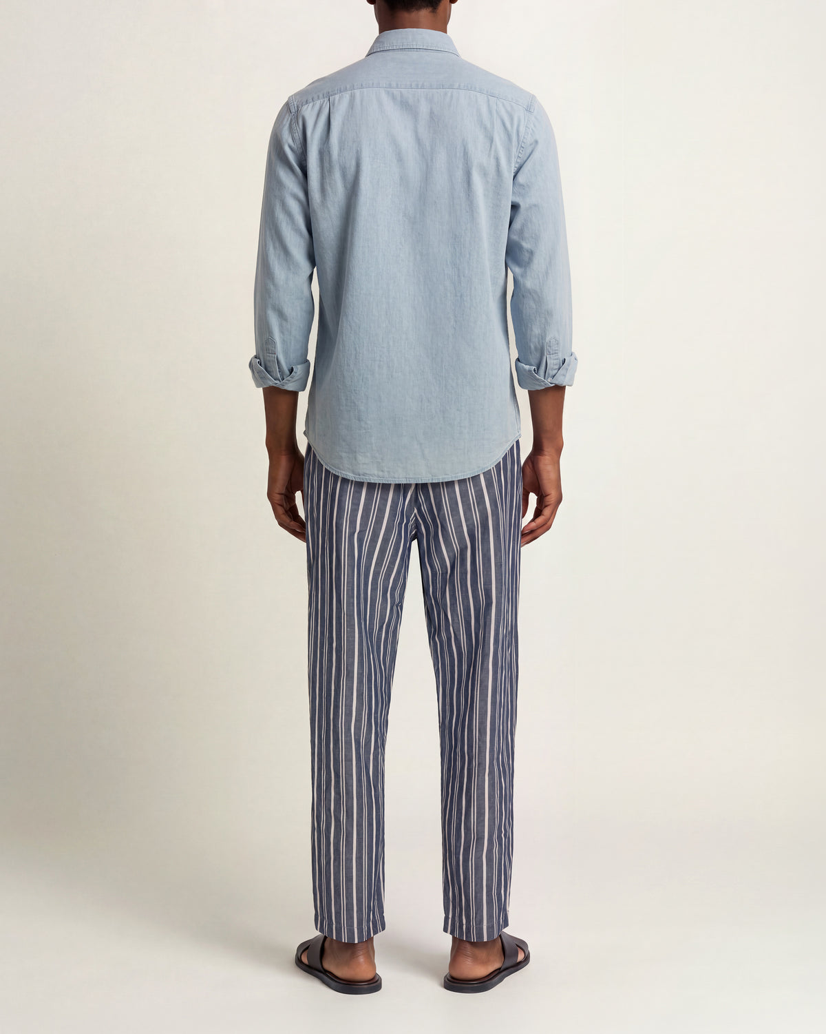 A man viewed from behind wearing a light blue long-sleeve button-up shirt and blue-and-white vertically striped trousers, paired with brown sandals, against a plain studio background.