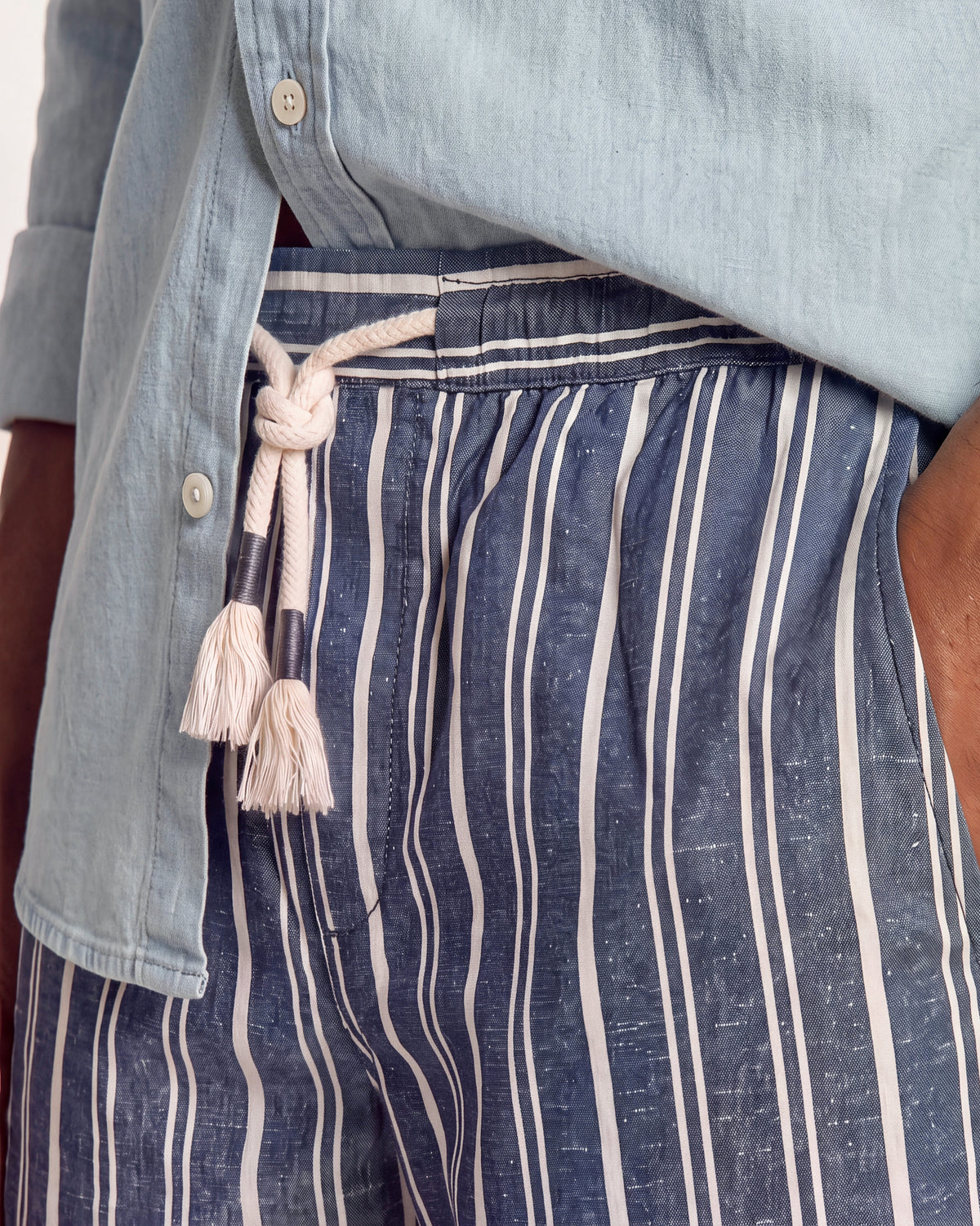 Close-up of a person wearing navy and white vertically striped shorts with a cream rope drawstring, paired with a light blue button-up shirt.