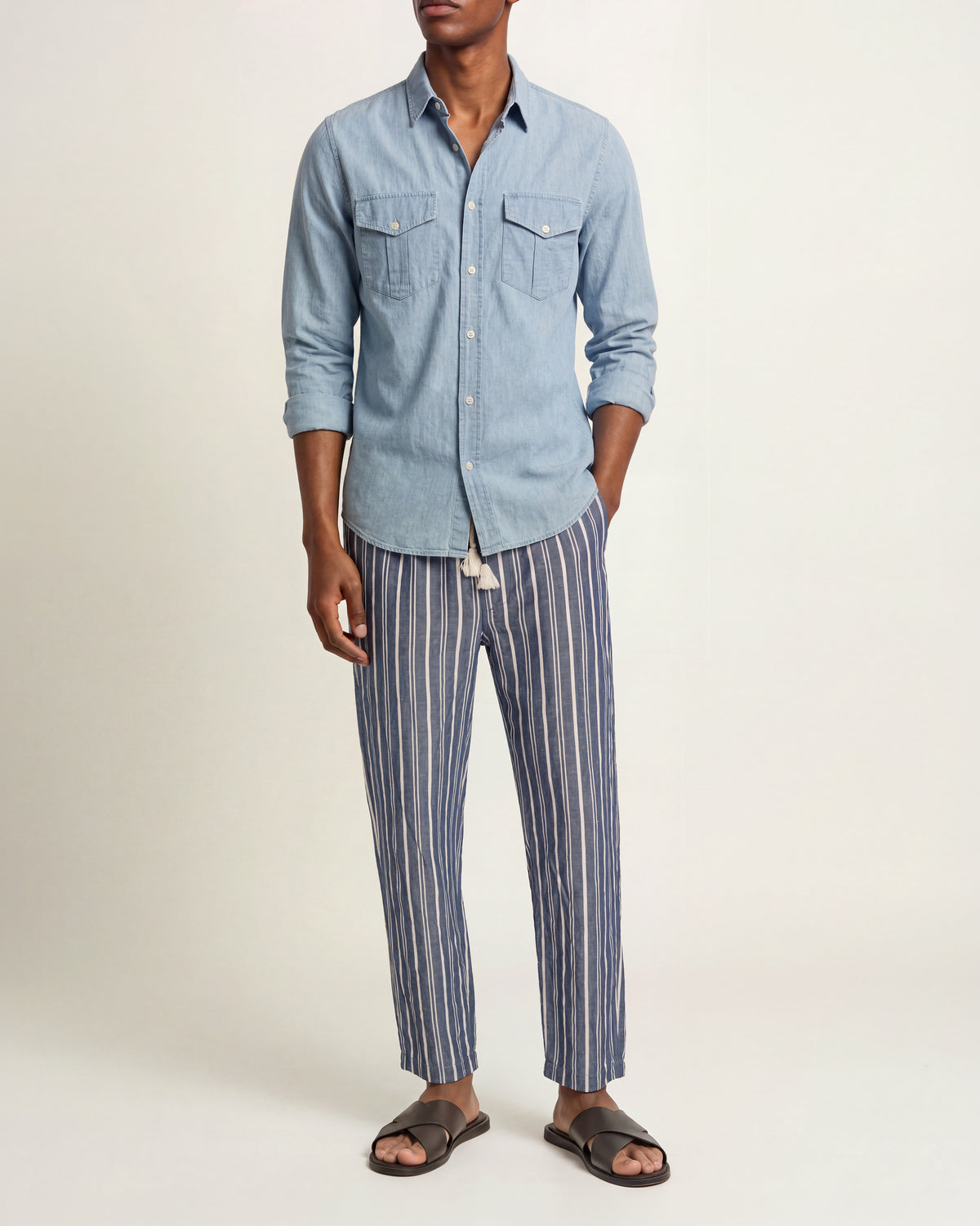 A man wearing a light blue long-sleeve button-up shirt and blue-and-white vertically striped trousers, paired with dark brown slide sandals, against a plain studio background.