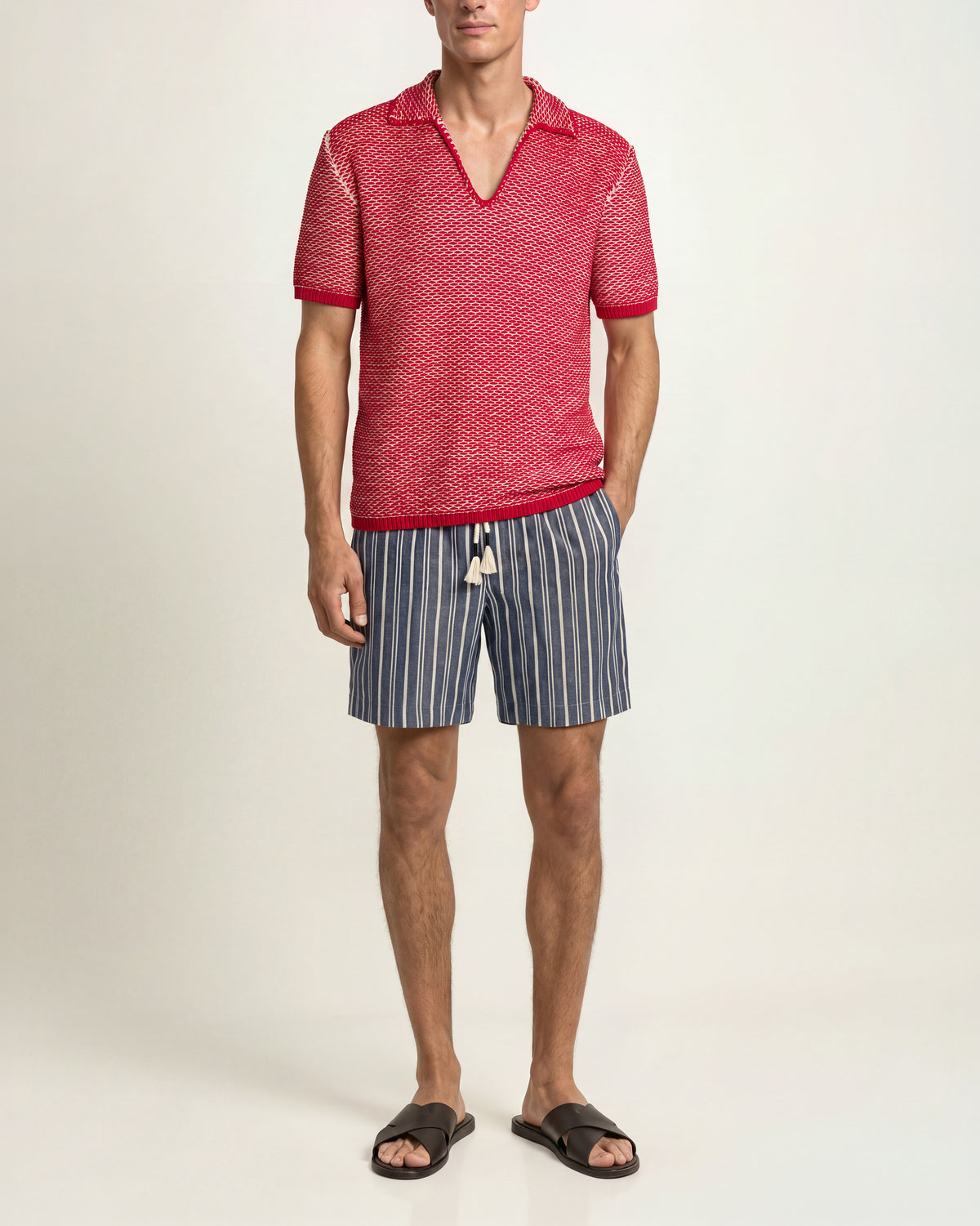 Man wearing a red-and-white striped short-sleeved crochet polo shirt, blue striped drawcord-waist shorts, and brown sandals on a plain background