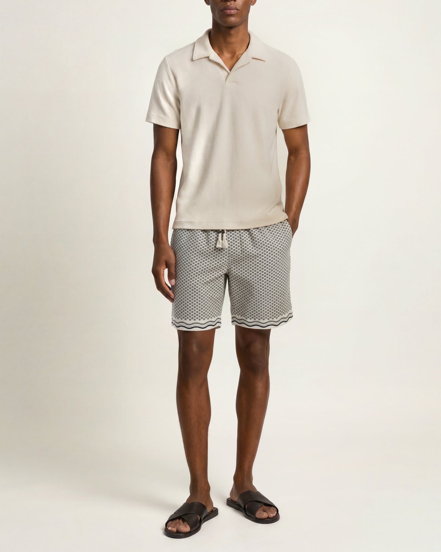 A man modeling a light beige short-sleeve polo shirt and black-and-white patterned shorts against a plain light beige studio background.