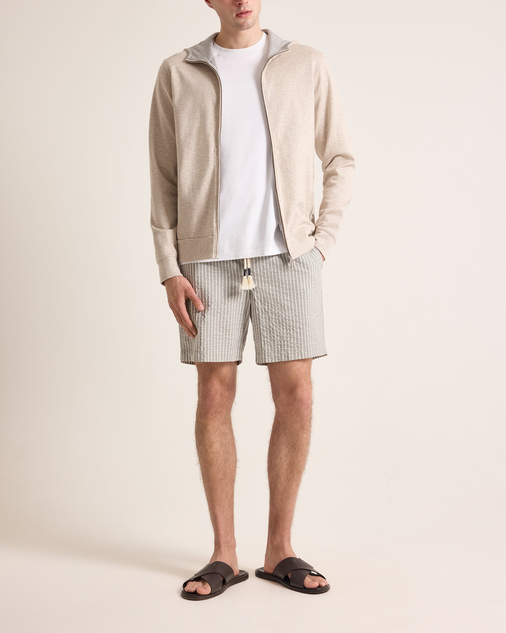 Man wearing a beige cardigan, white t-shirt, checkered shorts, and sandals on a plain background