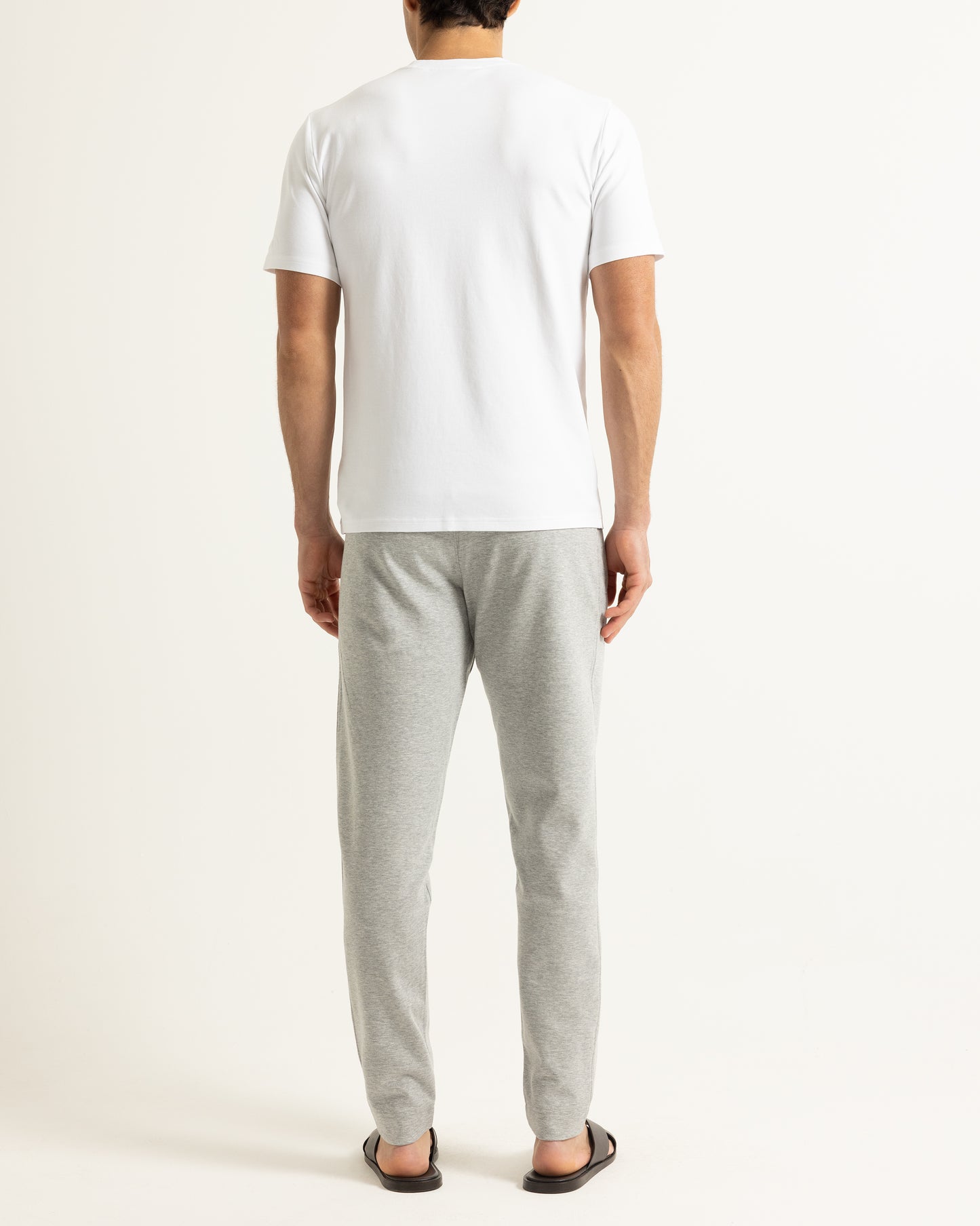 A man viewed from behind wearing a plain white short-sleeve t-shirt, light gray heather sweatpants, and dark brown sandals, standing against a solid off-white studio background.