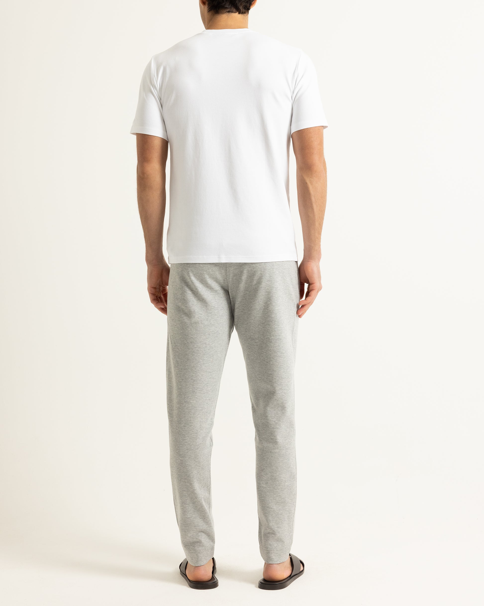 A man viewed from behind wearing a plain white short-sleeve t-shirt, light gray heather sweatpants, and dark brown sandals, standing against a solid off-white studio background.