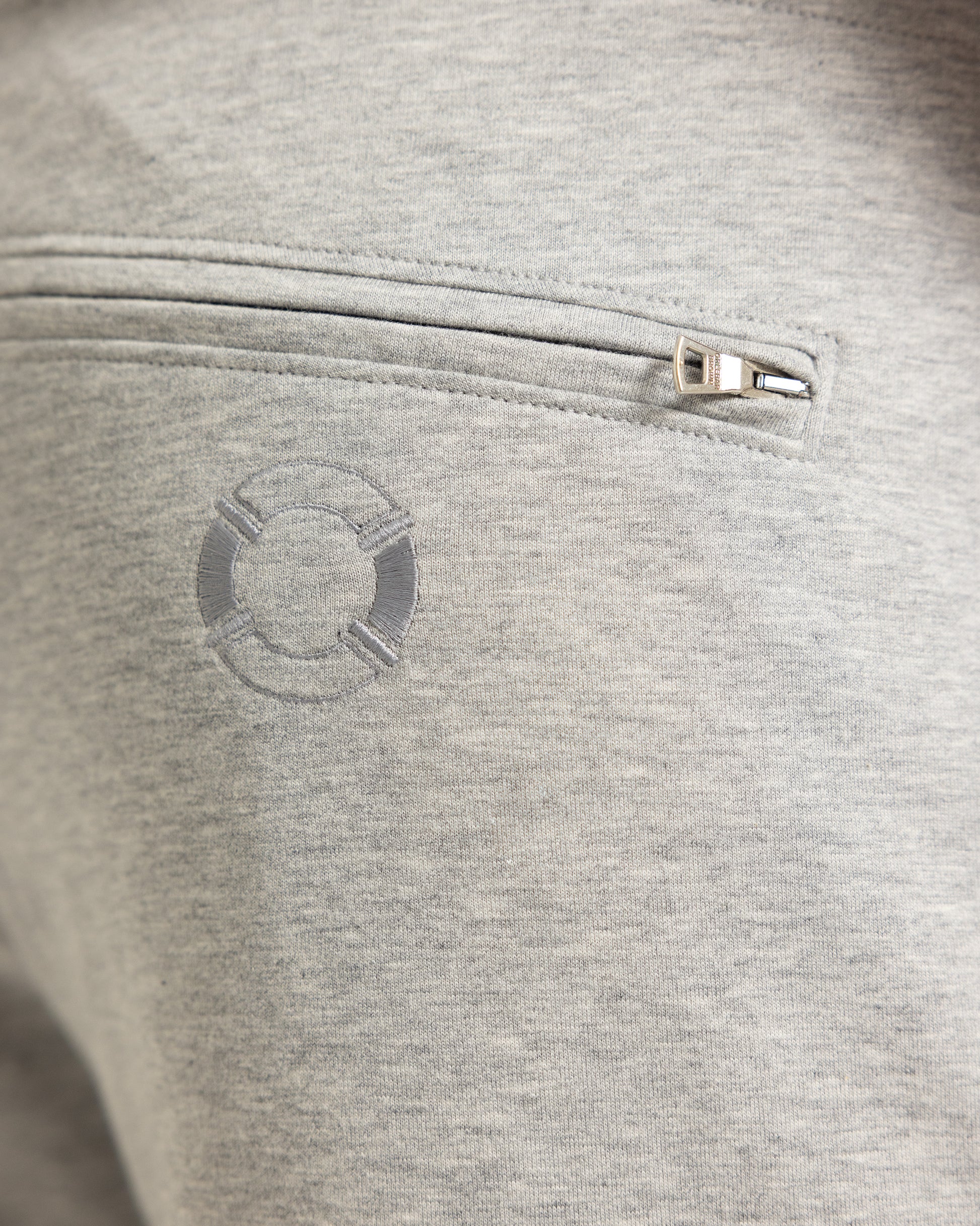 Close-up of a light heather gray fabric pocket with a silver zipper pull and a tonal circular logo on the front.