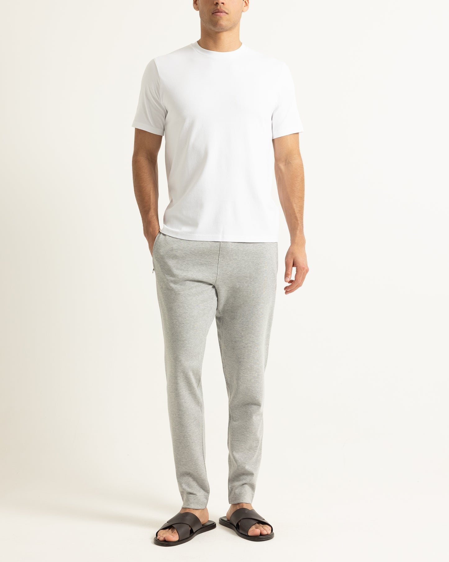 A man wearing a plain white short-sleeve t-shirt, light gray heather sweatpants, and black slide sandals, standing against a clean off-white studio background.