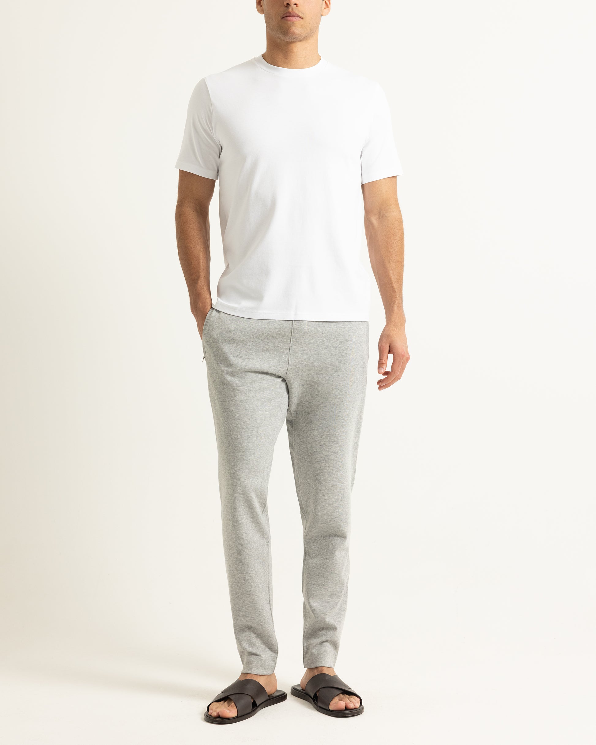 A man wearing a plain white short-sleeve t-shirt, light gray heather sweatpants, and black slide sandals, standing against a clean off-white studio background.