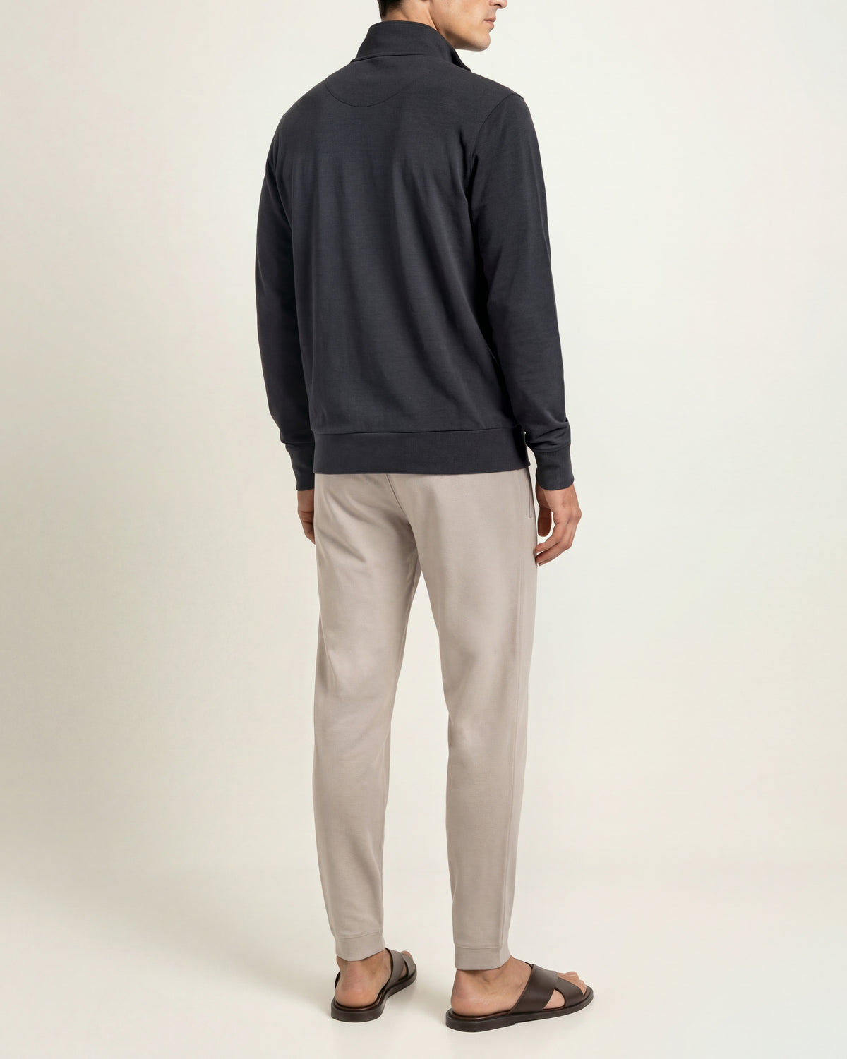 A man shown from the back wearing a dark charcoal gray quarter-zip sweatshirt, light beige trousers, and dark brown sandals against a plain off-white studio background.