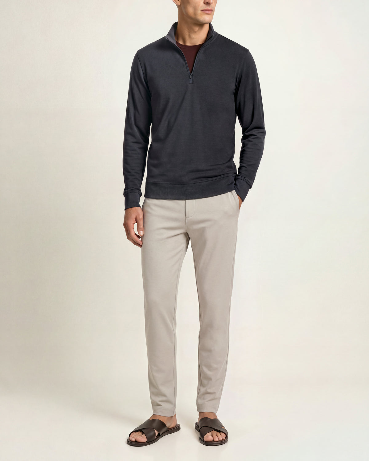 A man modeling a dark charcoal quarter-zip long-sleeve shirt, light beige trousers, and brown slide sandals against a plain off-white studio background.