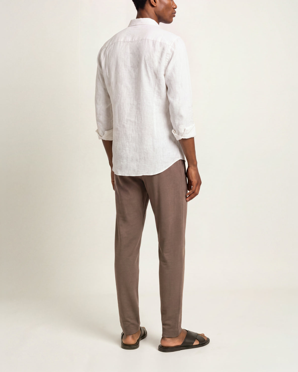 A man shown from the back wearing a white linen button-up shirt, brown trousers, and  brown slide sandals, standing against a plain white studio background.