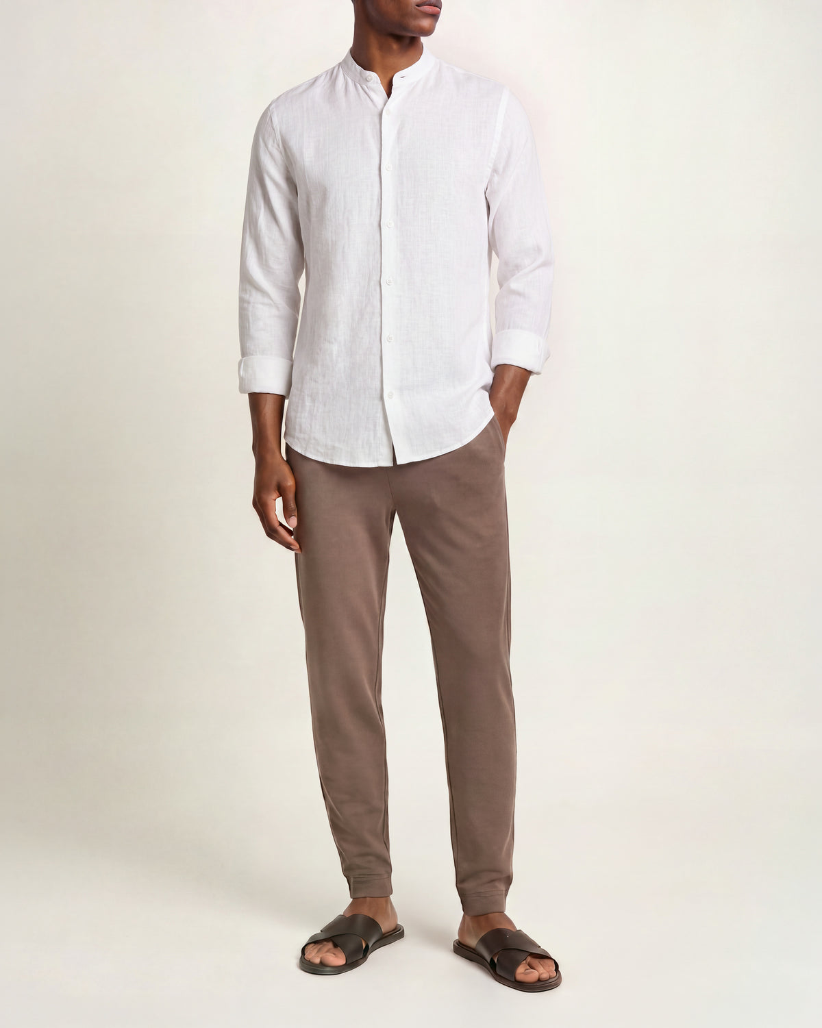 A man wearing a white long-sleeve linen shirt with a stand collar, paired with brown trousers and dark brown slide sandals, against a plain white studio background.