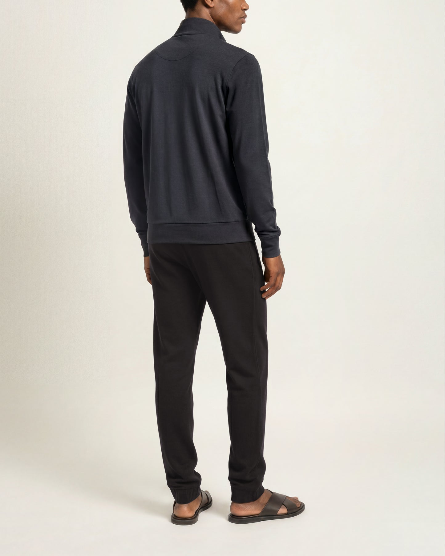 A man shown from the back wearing a dark navy quarter-zip sweatshirt and black sweatpants, standing against a plain off-white studio background.