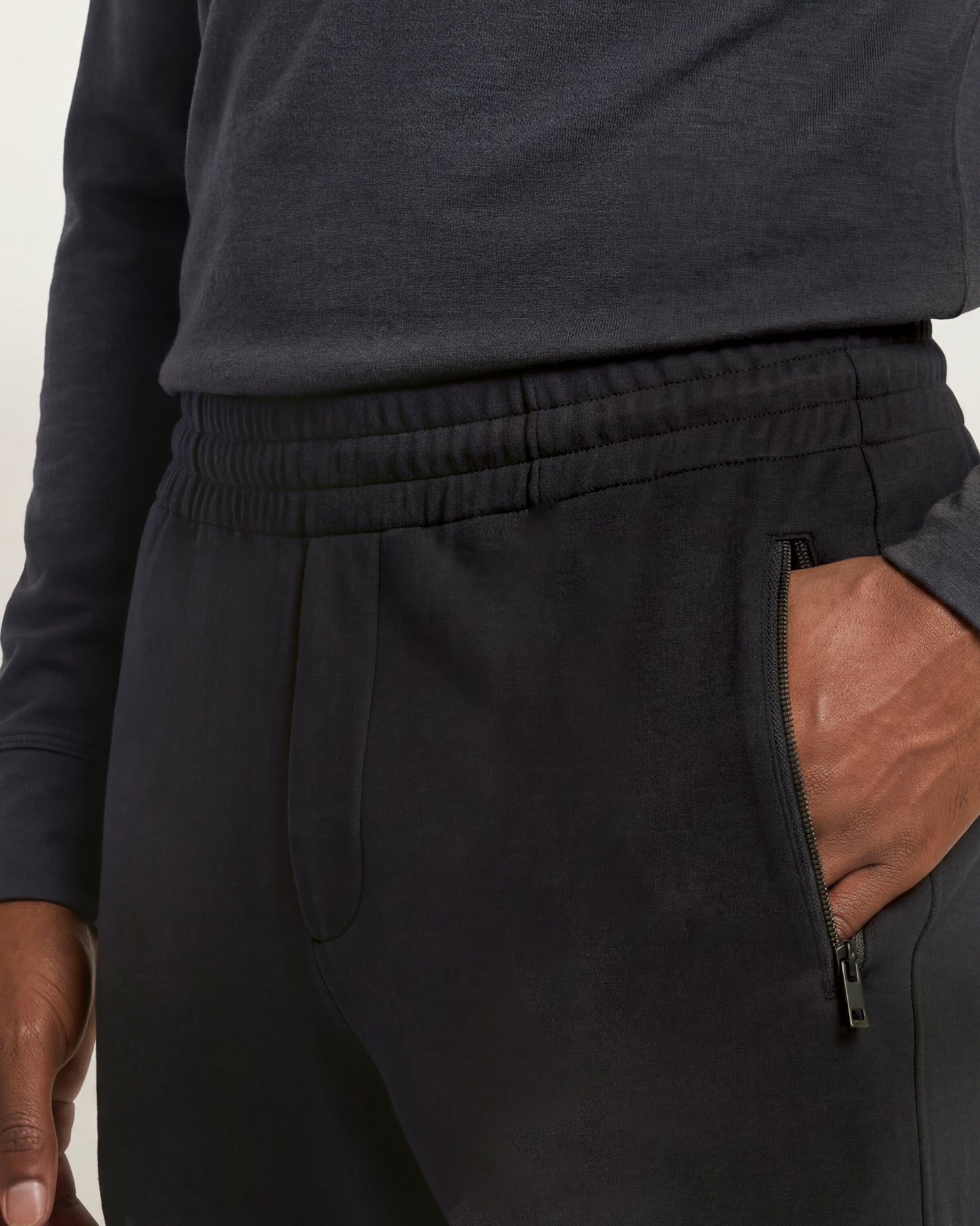 Close-up of a person wearing black sweatpants with an elastic waistband, side zip pocket, and hand in pocket, paired with a dark charcoal long-sleeve top.
