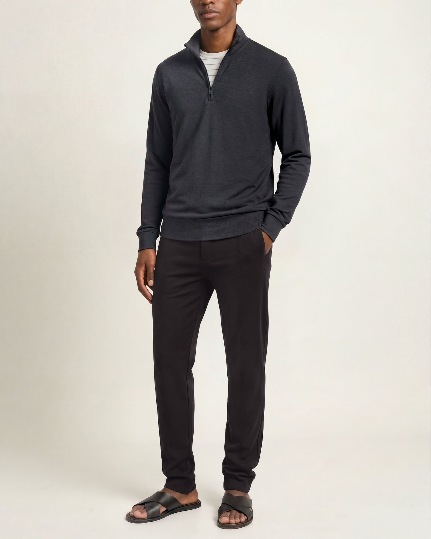 A man modeling a dark charcoal gray quarter-zip pullover sweater, black pants, and dark sandals against a plain off-white studio background.
