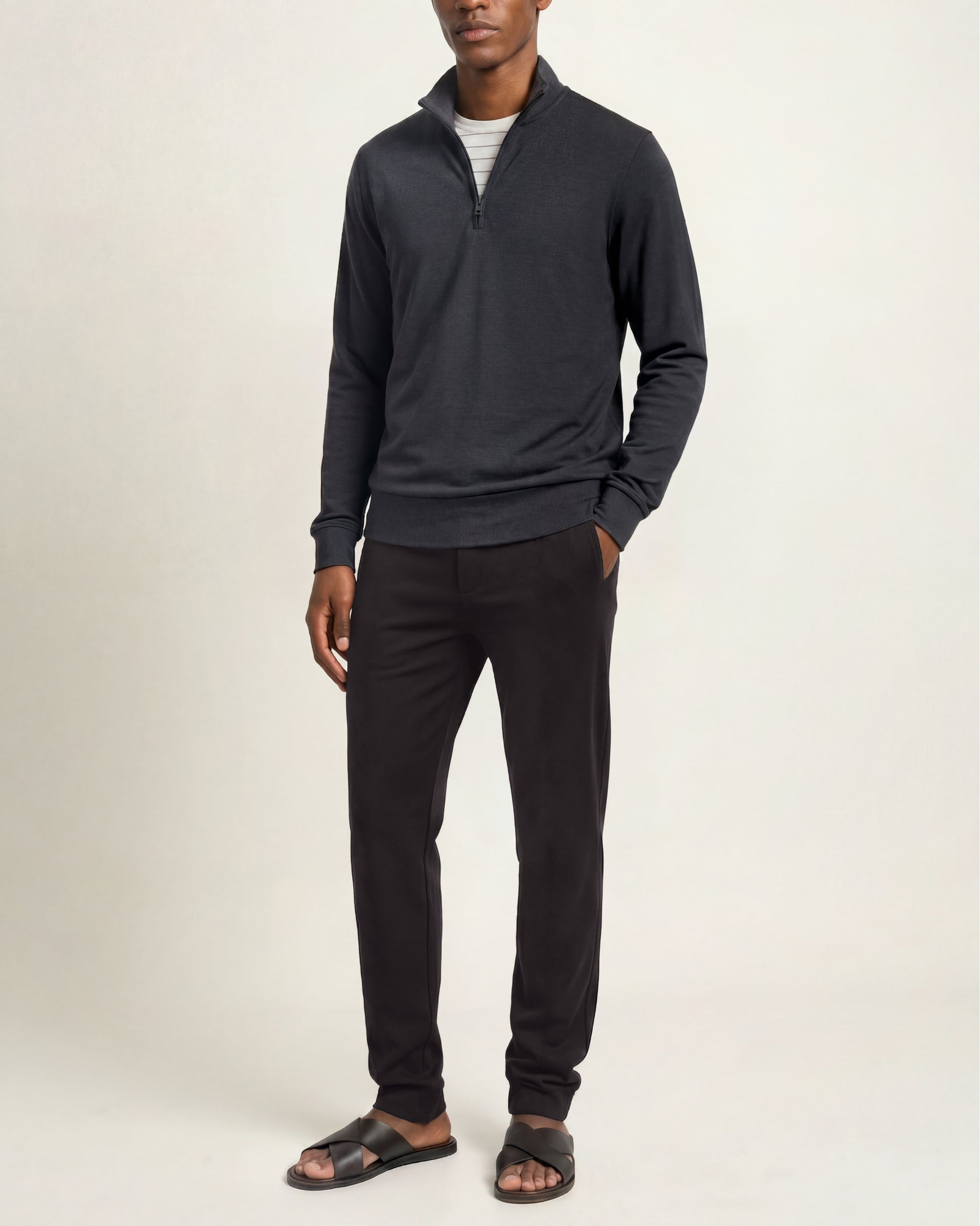 A man modeling a dark charcoal gray quarter-zip pullover sweater, black pants, and dark sandals against a plain off-white studio background.