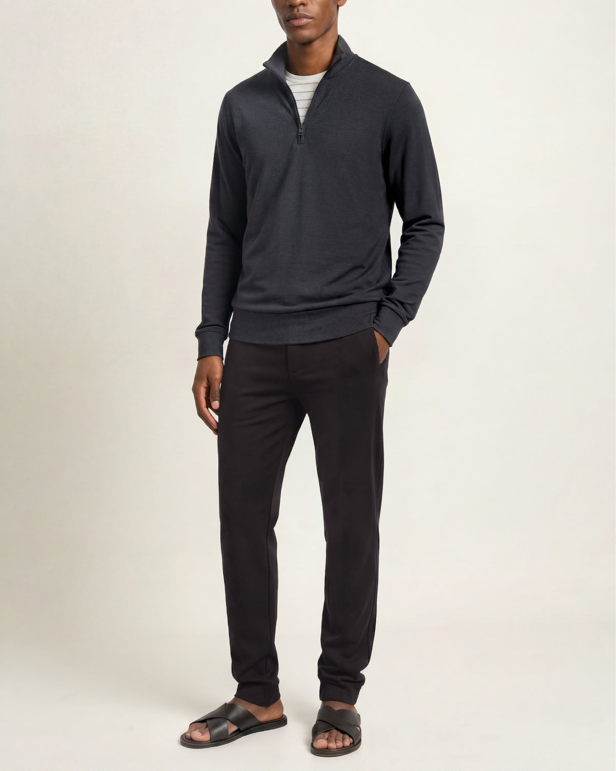 A man modeling a dark charcoal gray quarter-zip pullover sweater, black pants, and dark sandals against a plain off-white studio background.