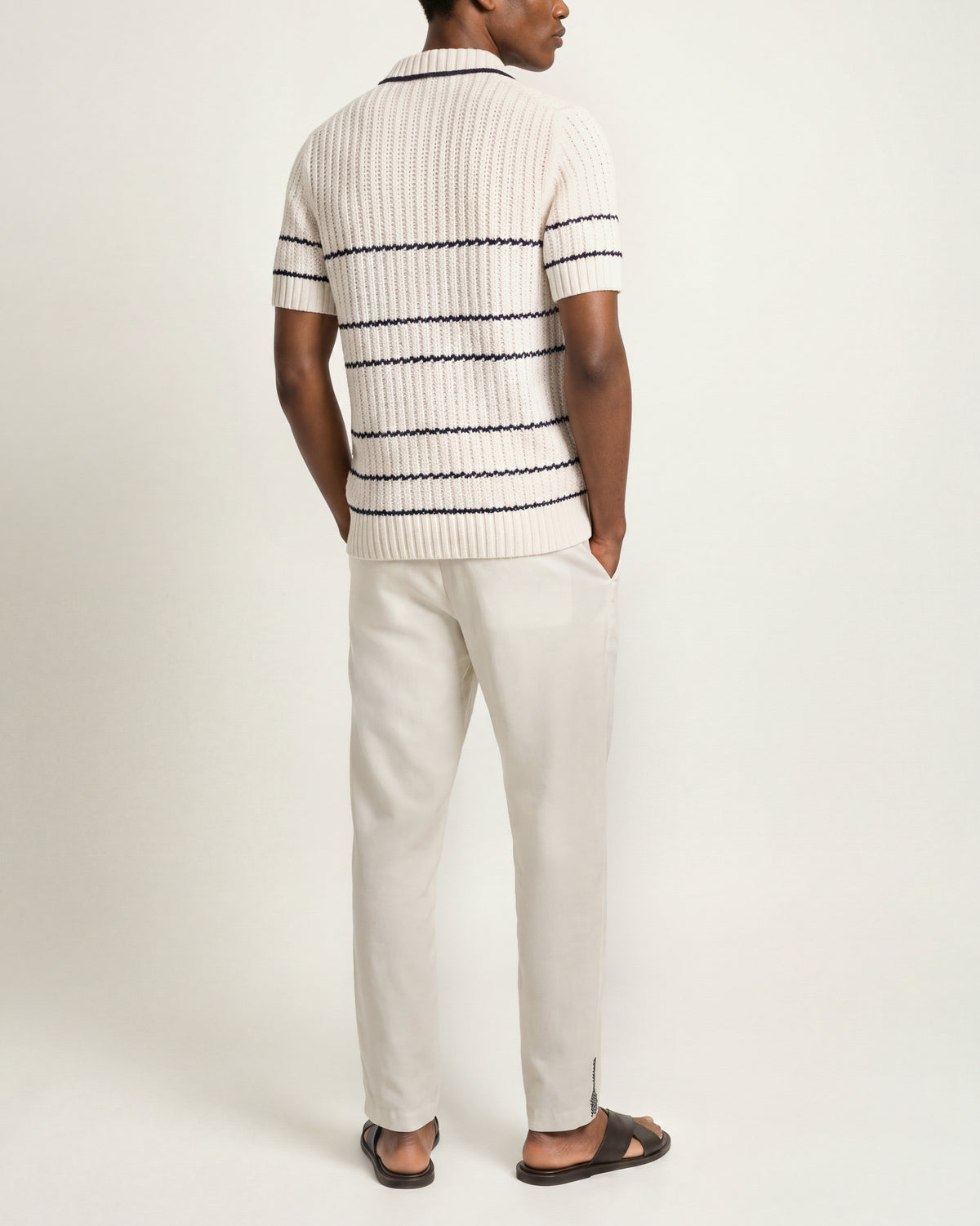 Man wearing white knitted polo shirt with black stripes detailing and white trousers on a plain background