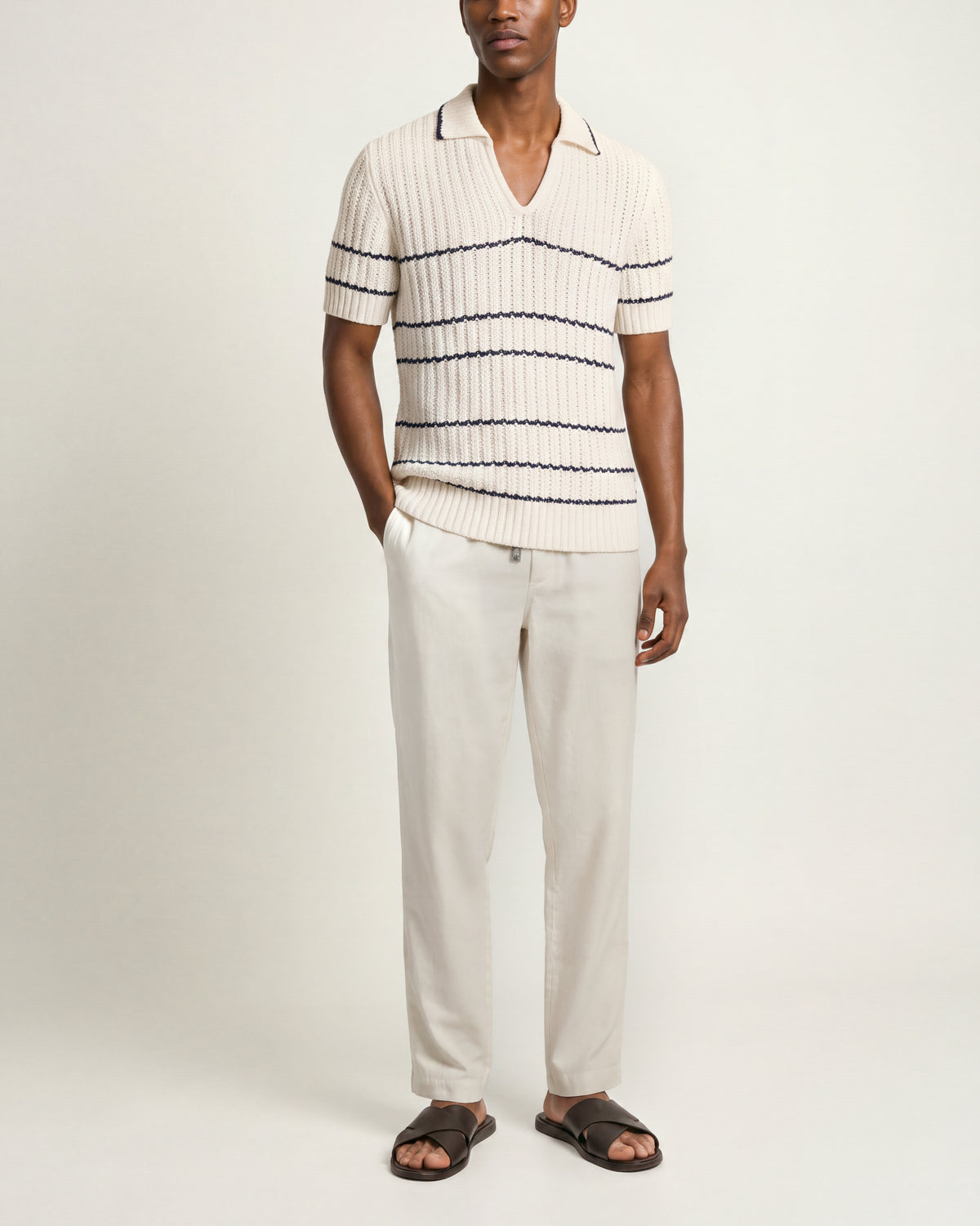 Man wearing white knitted polo shirt with black stripes detailing and white trousers on a plain background