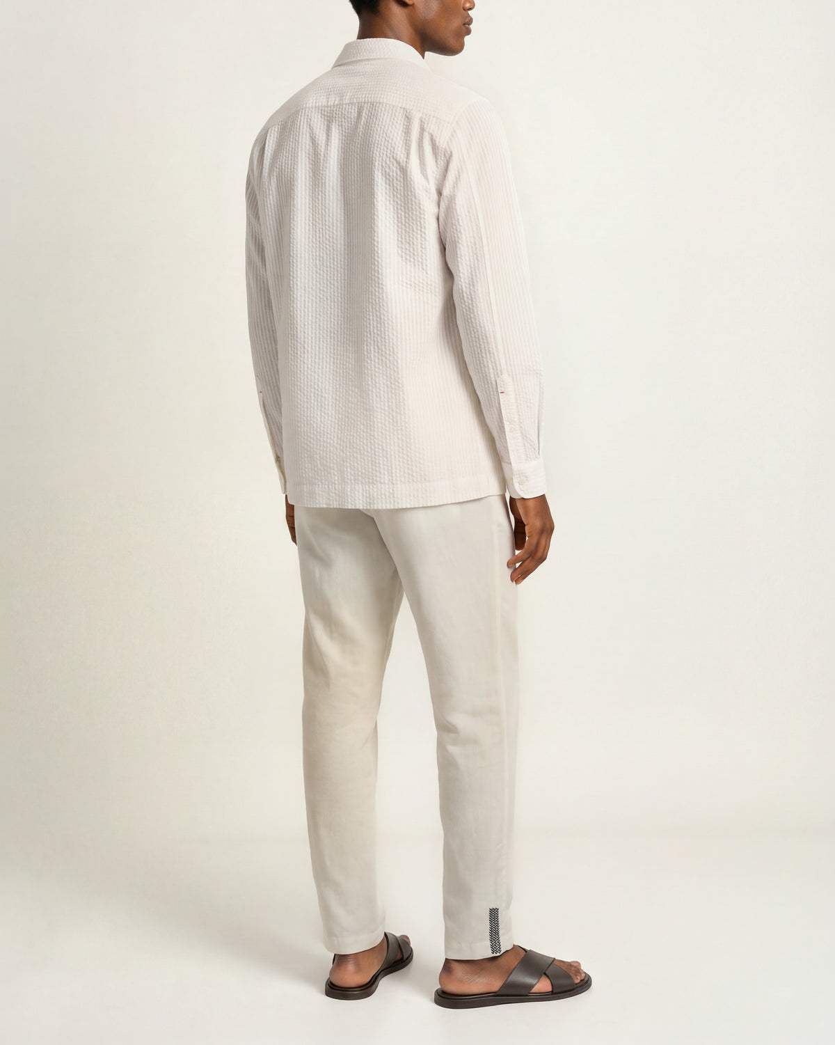 Back view of a man wearing white long-sleeved seersucker shirt with white drawcord waist trousers and brown sandals on a plain background