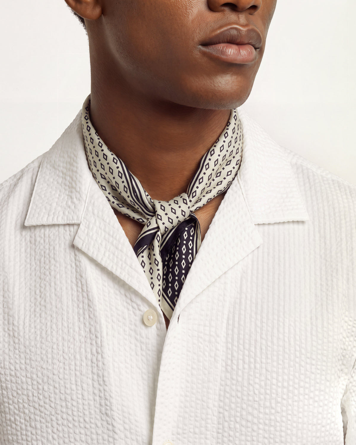 Closeup of man wearing white seersucker shirt with black and white patterned neck scarf on a plain background