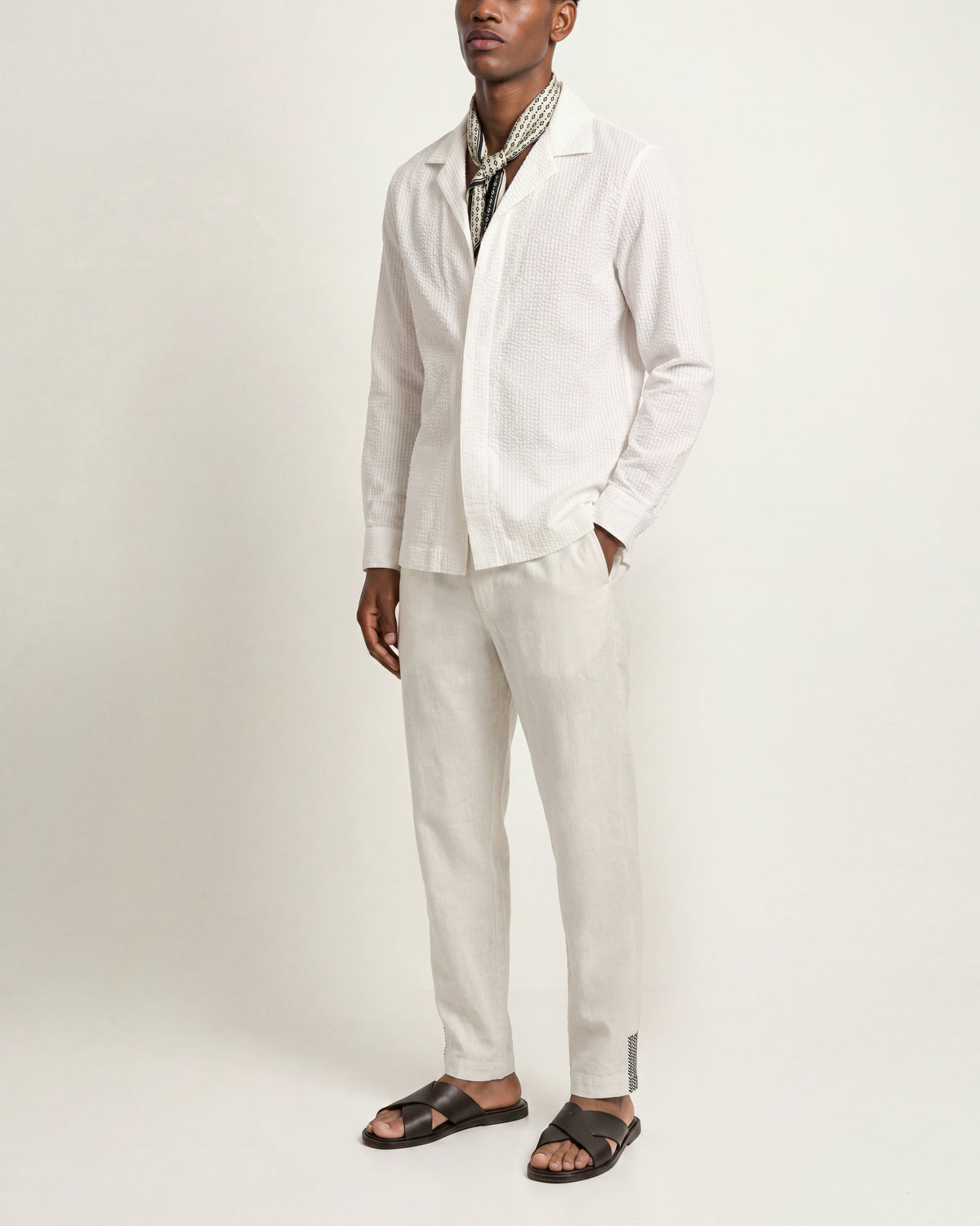Man wearing white long-sleeved shirt with patterned scarf paired with white drawcord waist trousers and black sandals on a plain background