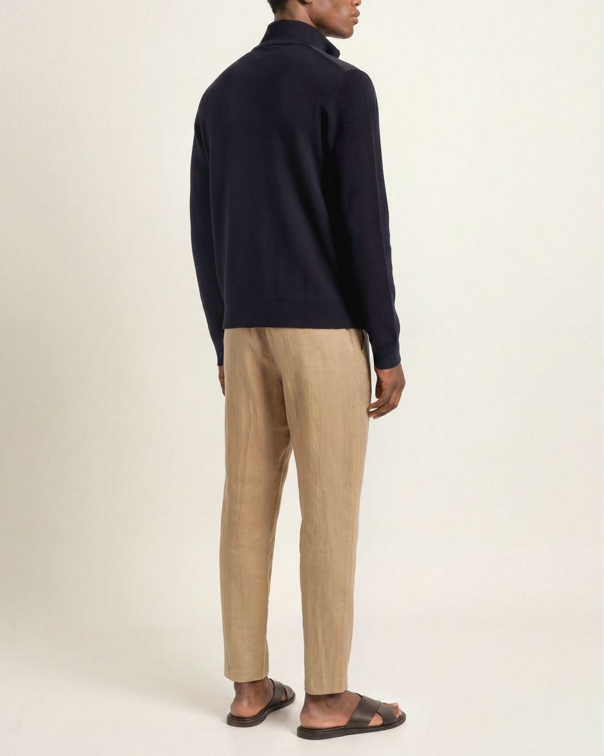 A man shown from the back wearing a dark navy knit quarter-zip sweater, beige trousers, and dark brown sandals, photographed against a plain light beige studio background.