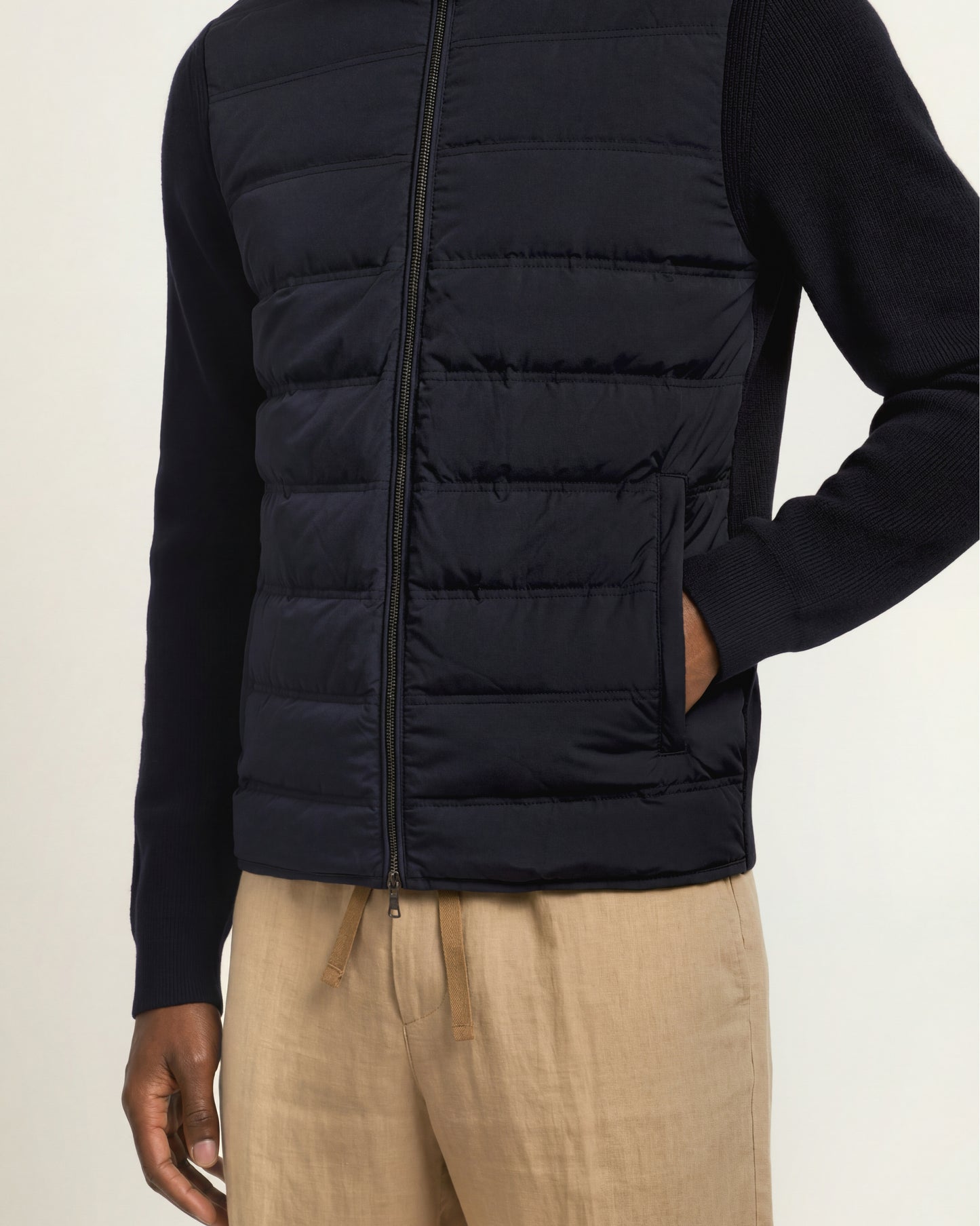 A man wearing a dark navy blue zip-up puffer vest over a matching long-sleeve knit top, paired with beige drawstring trousers, shown against a plain light background.
