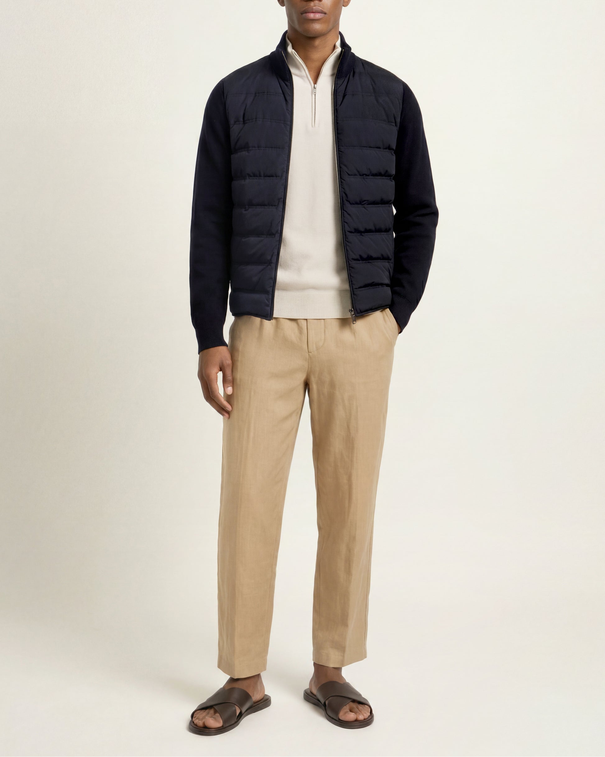 A man modeling a dark navy quilted puffer jacket over a cream zip-up sweater, paired with beige trousers and dark brown slide sandals, against a plain off-white studio background.