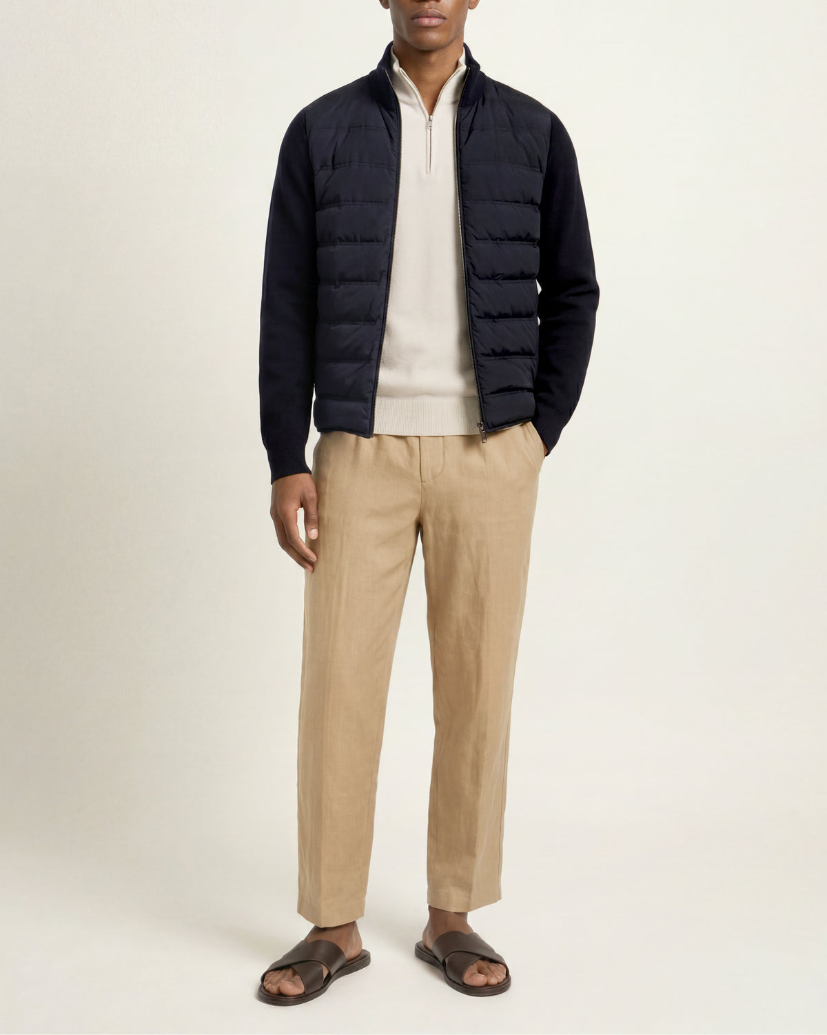A man modeling a dark navy quilted puffer jacket over a cream zip-up sweater, paired with beige trousers and dark brown slide sandals, against a plain off-white studio background.