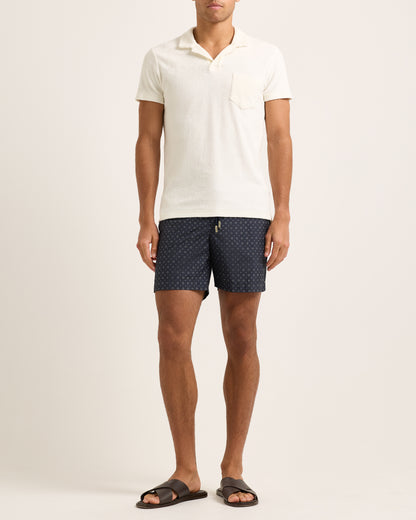 Person wearing a white towelling polo shirt and navy patterned shorts on a plain background