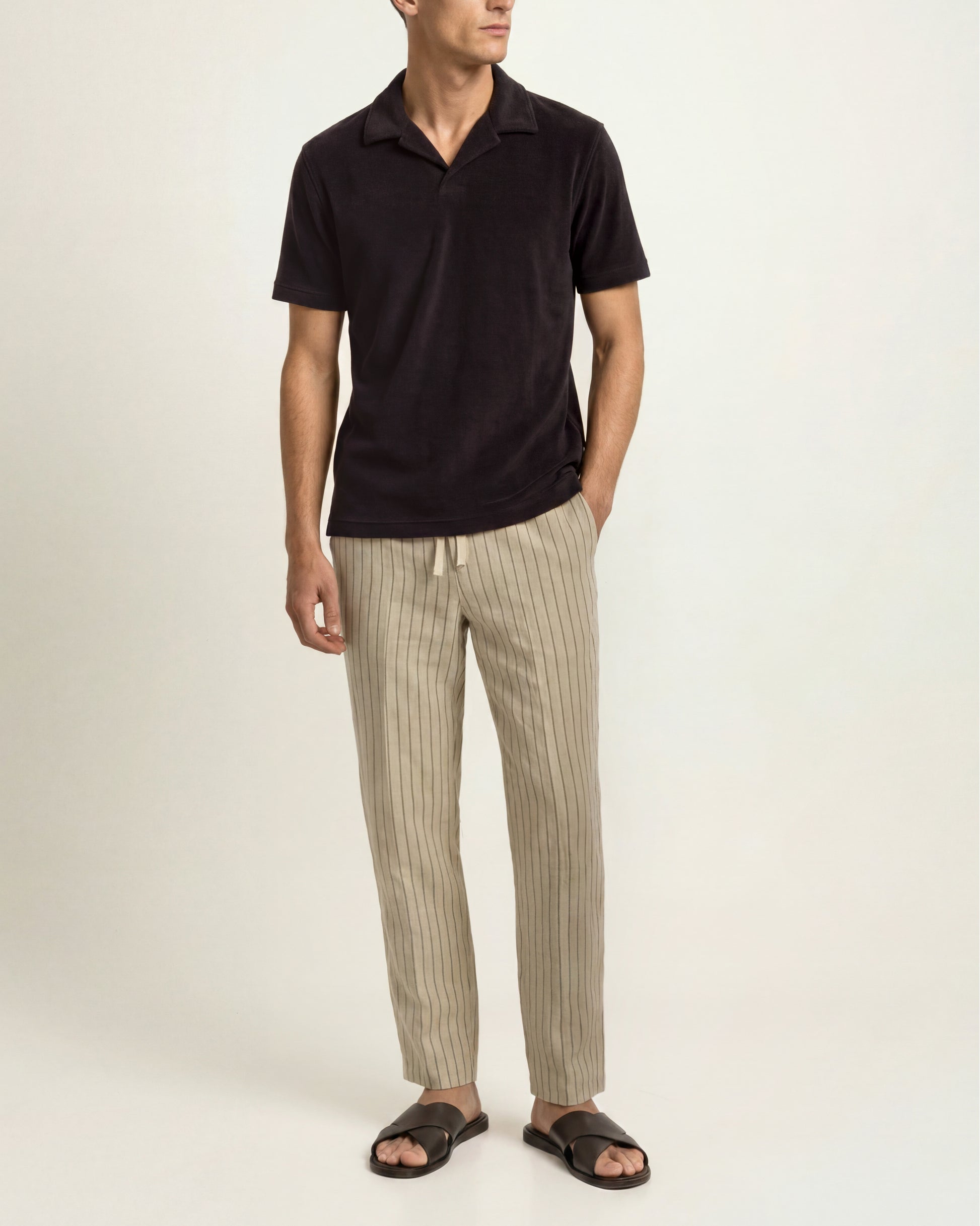 A man modeling a black short-sleeve polo shirt paired with beige pinstriped trousers and black slide sandals, standing against a plain off-white studio background.