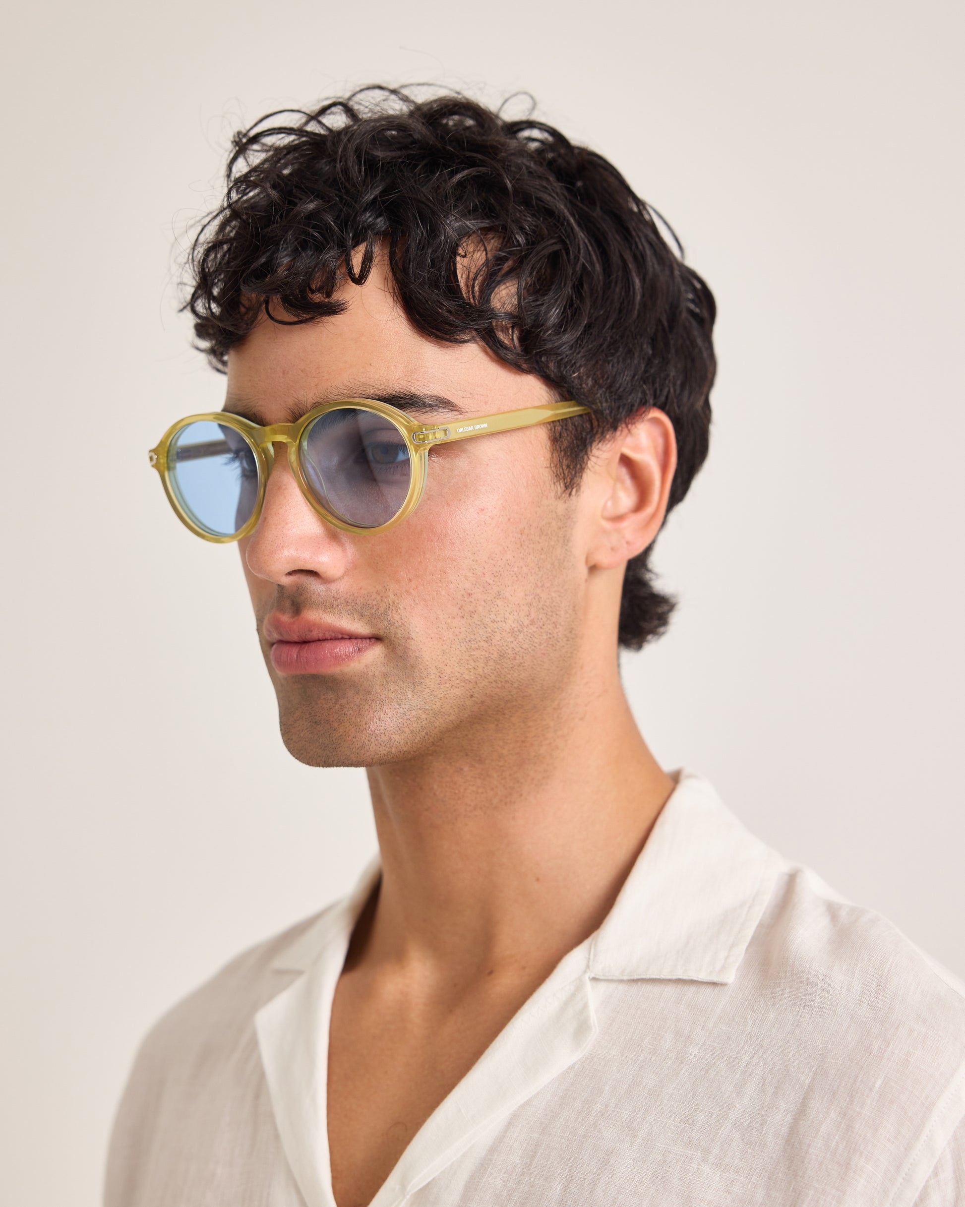 Man wearing gold-framed sunglasses and a white shirt against a plain background