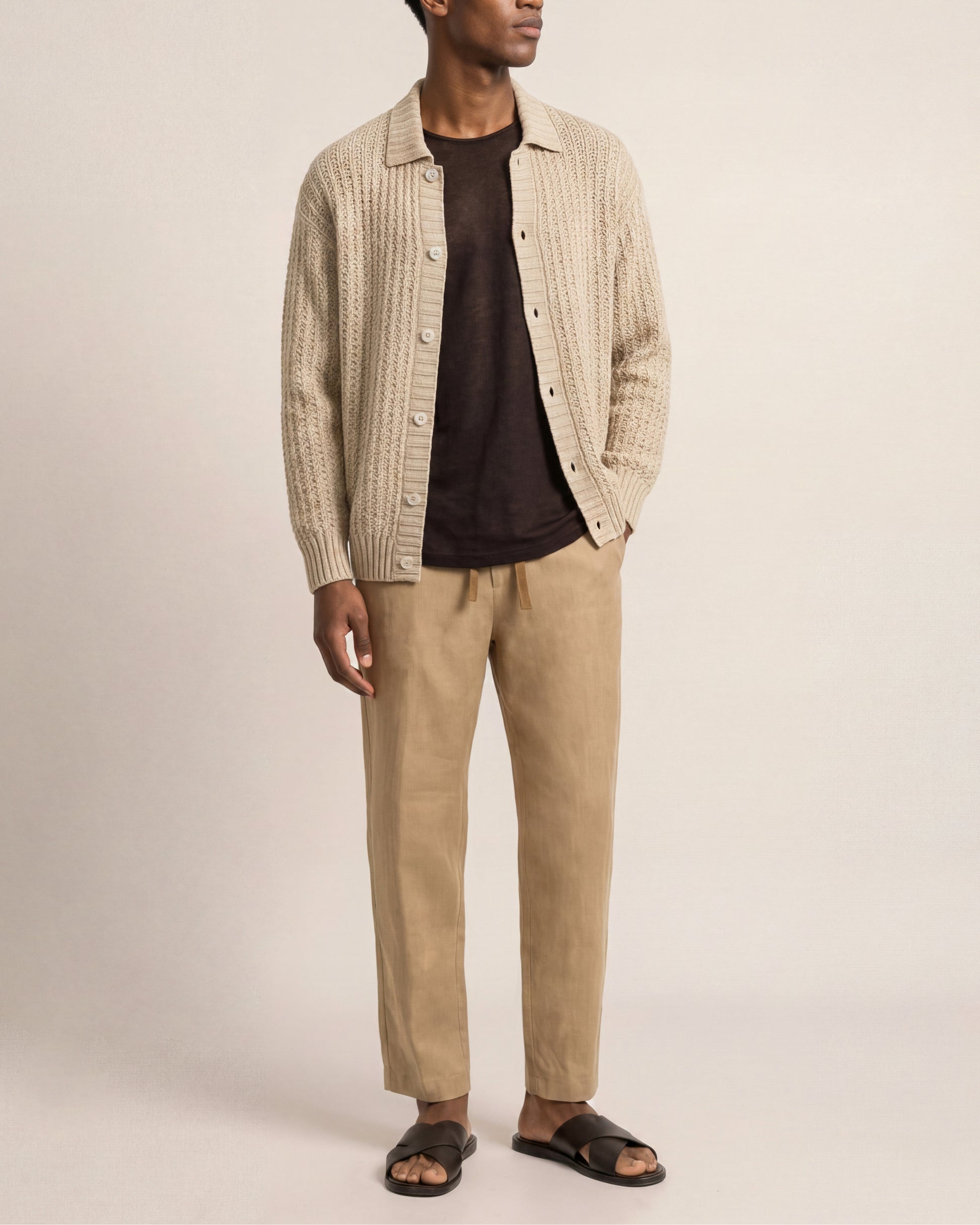 Man wearing a beige knitted shirt over a black t-shirt with beige pants on a plain background