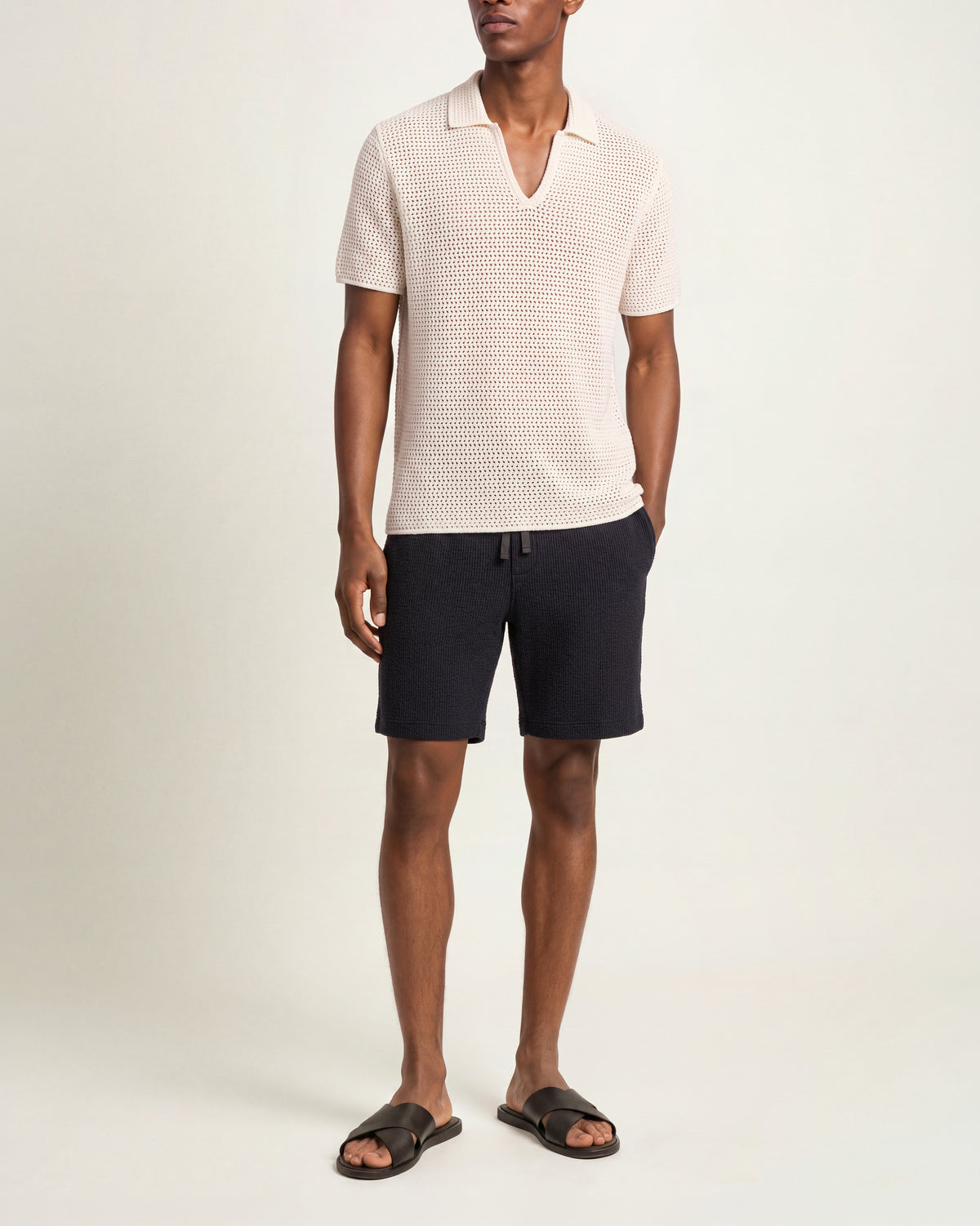 Man wearing white crochet stitch polo shirt with towelling sweat shorts and brown sandals on a plain background