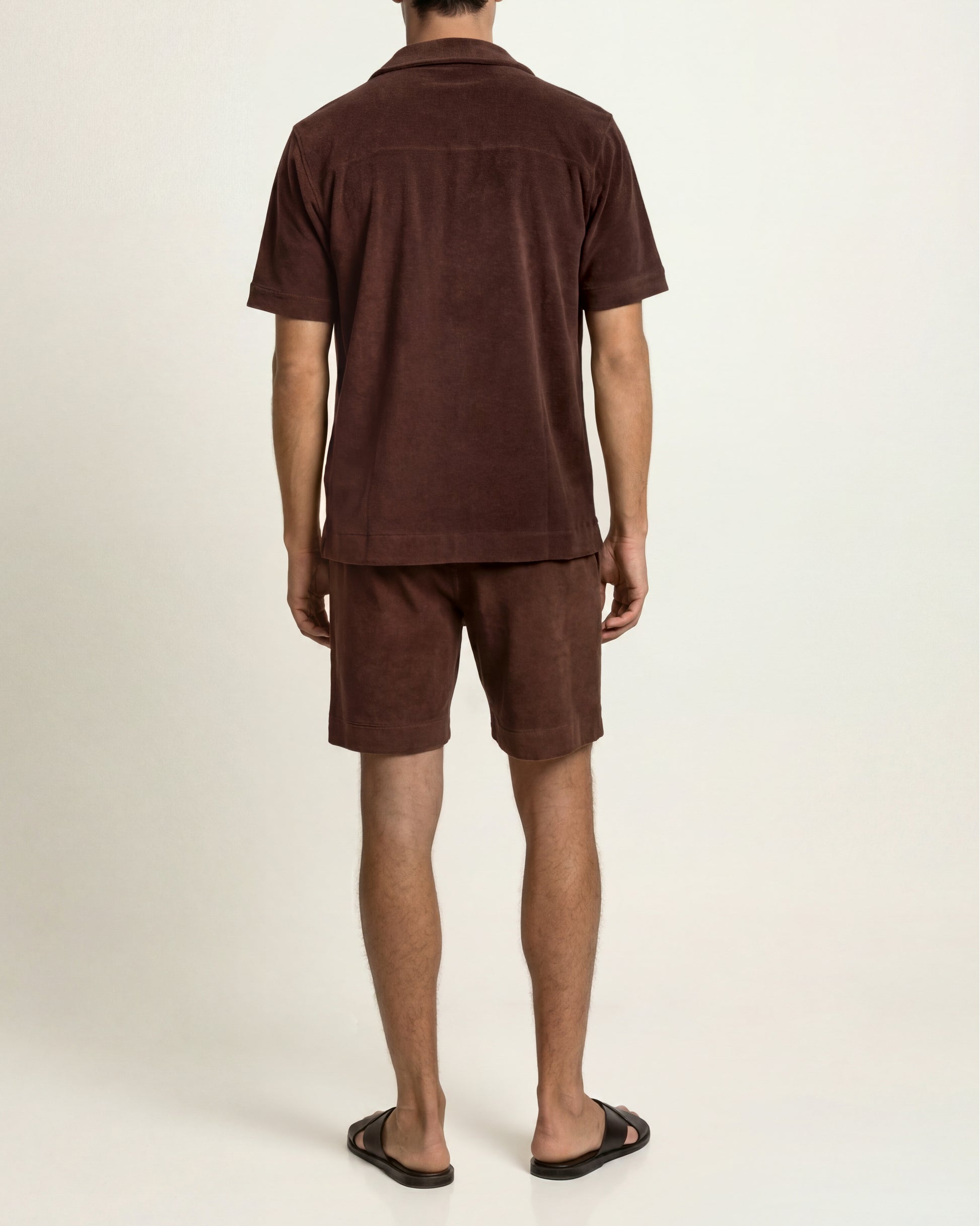 A man viewed from the back wearing a matching dark brown short-sleeve button-up shirt and shorts set, paired with black slide sandals, against a plain off-white studio background.