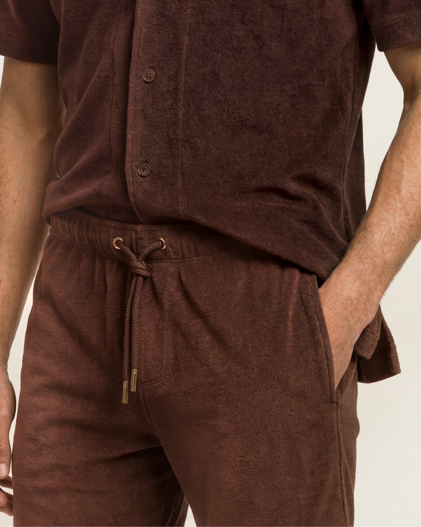 A close-up of a person wearing a matching brown lounge set made of textured fabric, featuring a short-sleeve button-front top and drawstring pants.