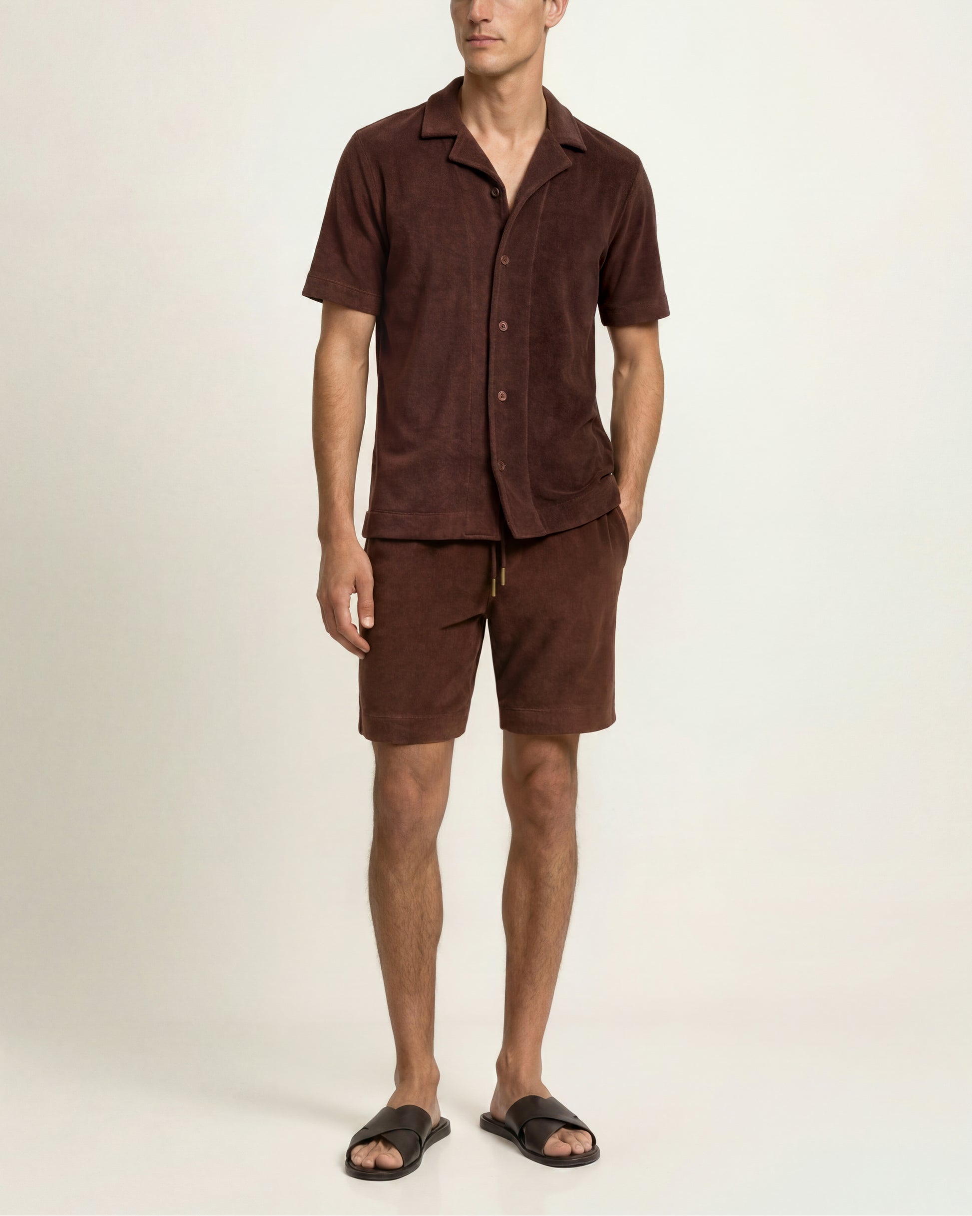 A man modeling a matching brown short-sleeve button-up shirt and shorts set, paired with black slide sandals, against a plain off-white studio background.