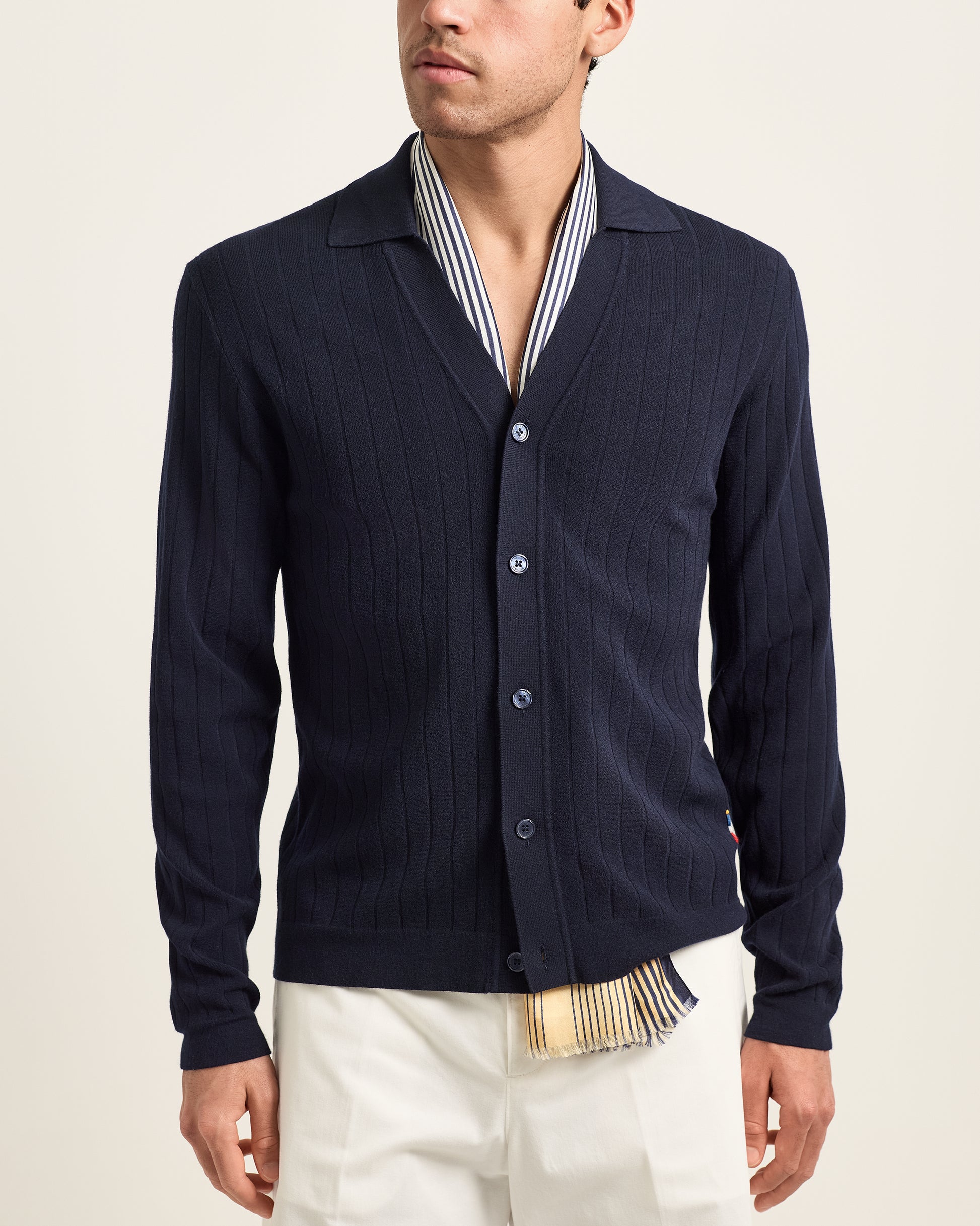 Man wearing a navy cardigan over a striped shirt with a beige background