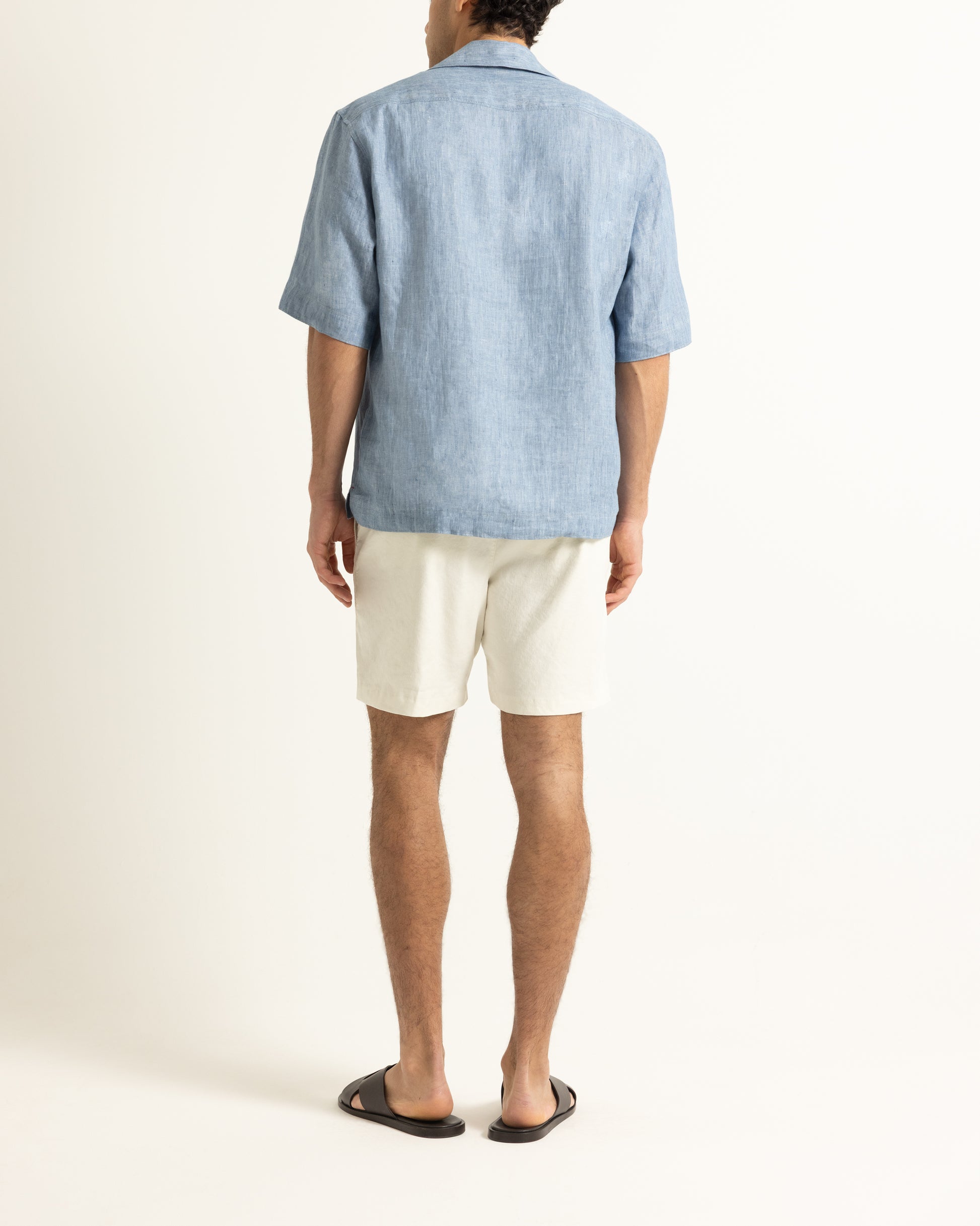 Person wearing a light blue linen shirt and white shorts on a plain background